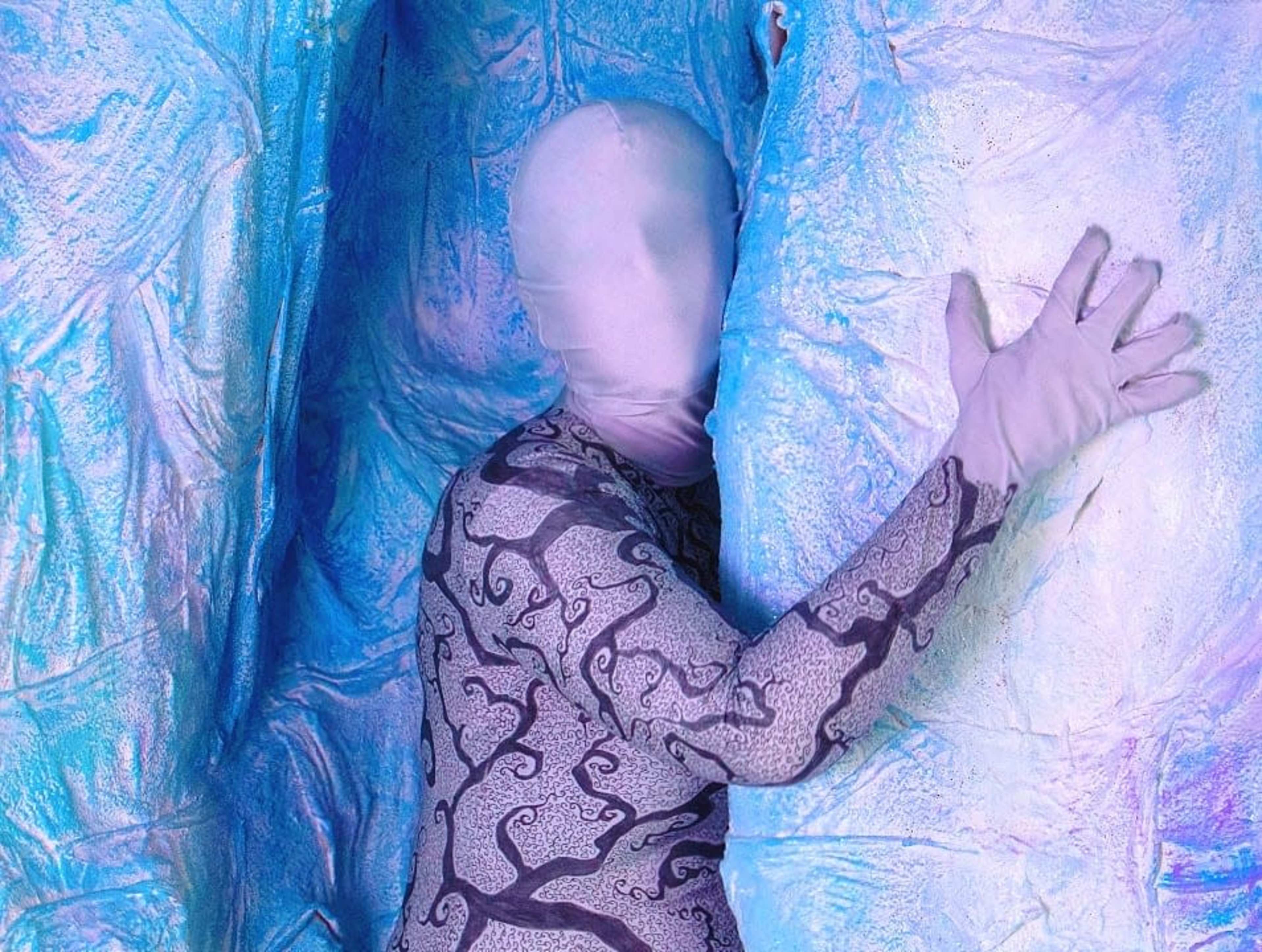 A figure in a patterned bodysuit and matching gloves poses against a textured, blue and silver background.