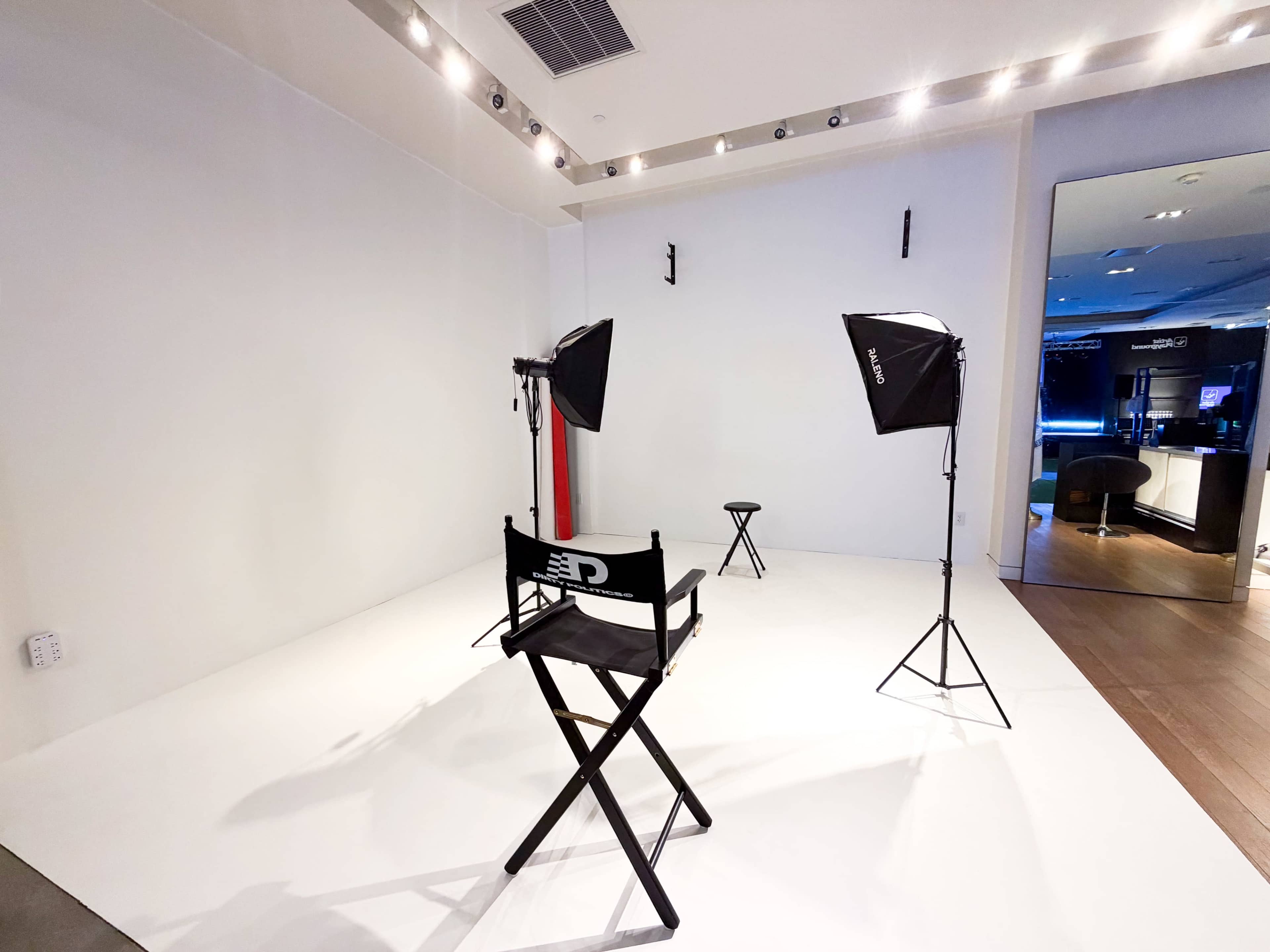 The image shows a minimalist photo studio with a white backdrop, a director's chair, and two lighting setups.
