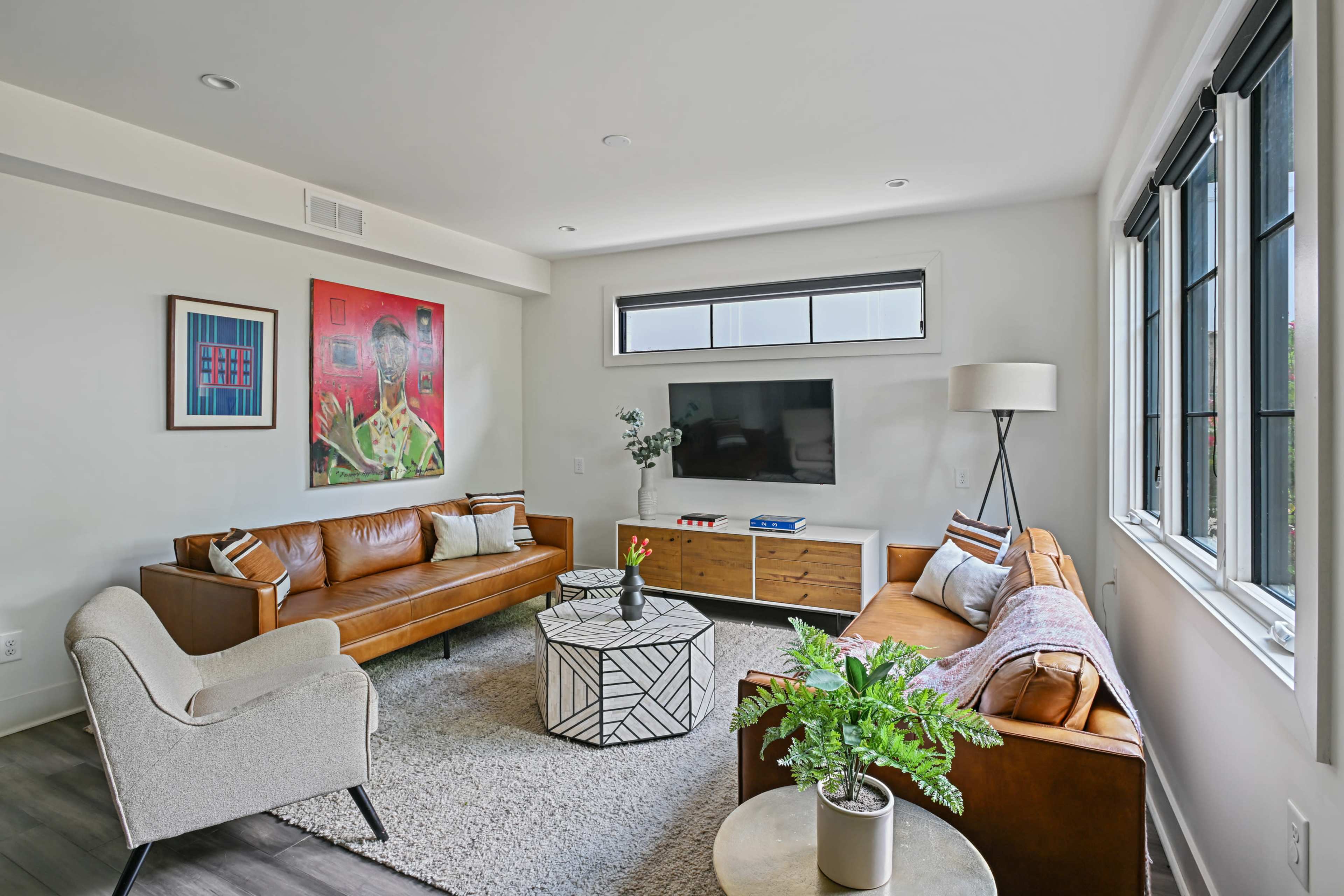 The image shows a modern living room with two sofas, a round coffee table, a wall-mounted television, and a large artwork above the brown leather couch.