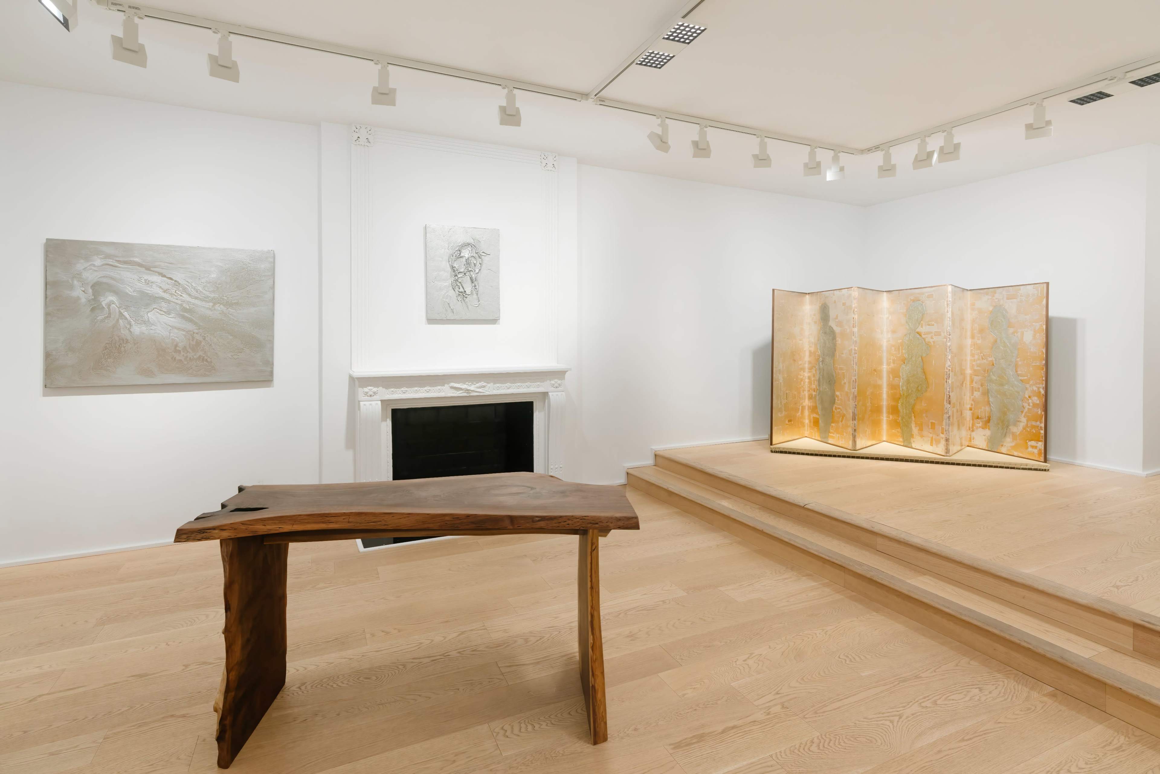 An art gallery with a wooden table, a folding screen, and two framed artworks displayed on white walls.