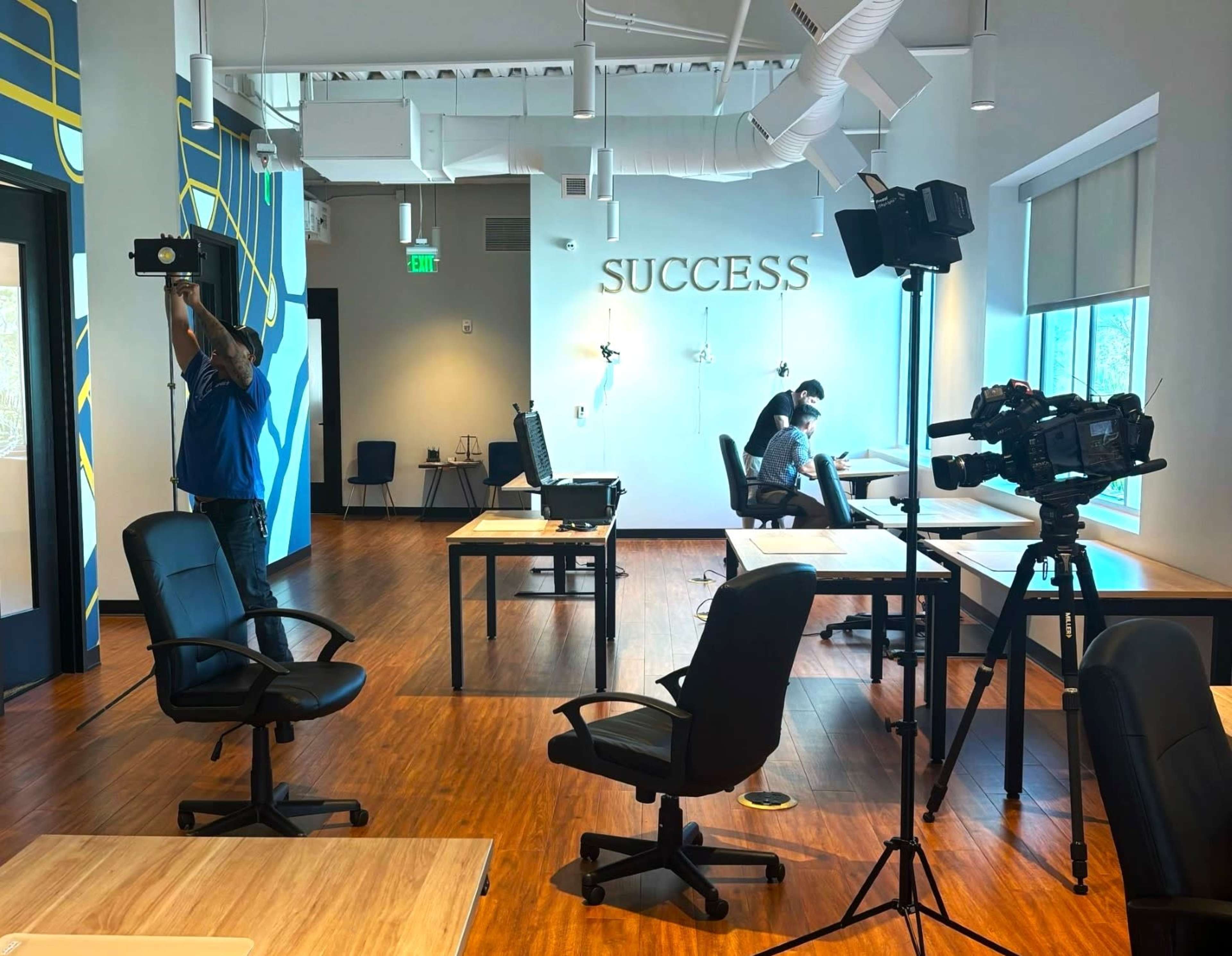 A man adjusts a light above a desk in an office setting with a large "SUCCESS" sign on the wall, while another person works at a nearby desk with cameras set up in the foreground.