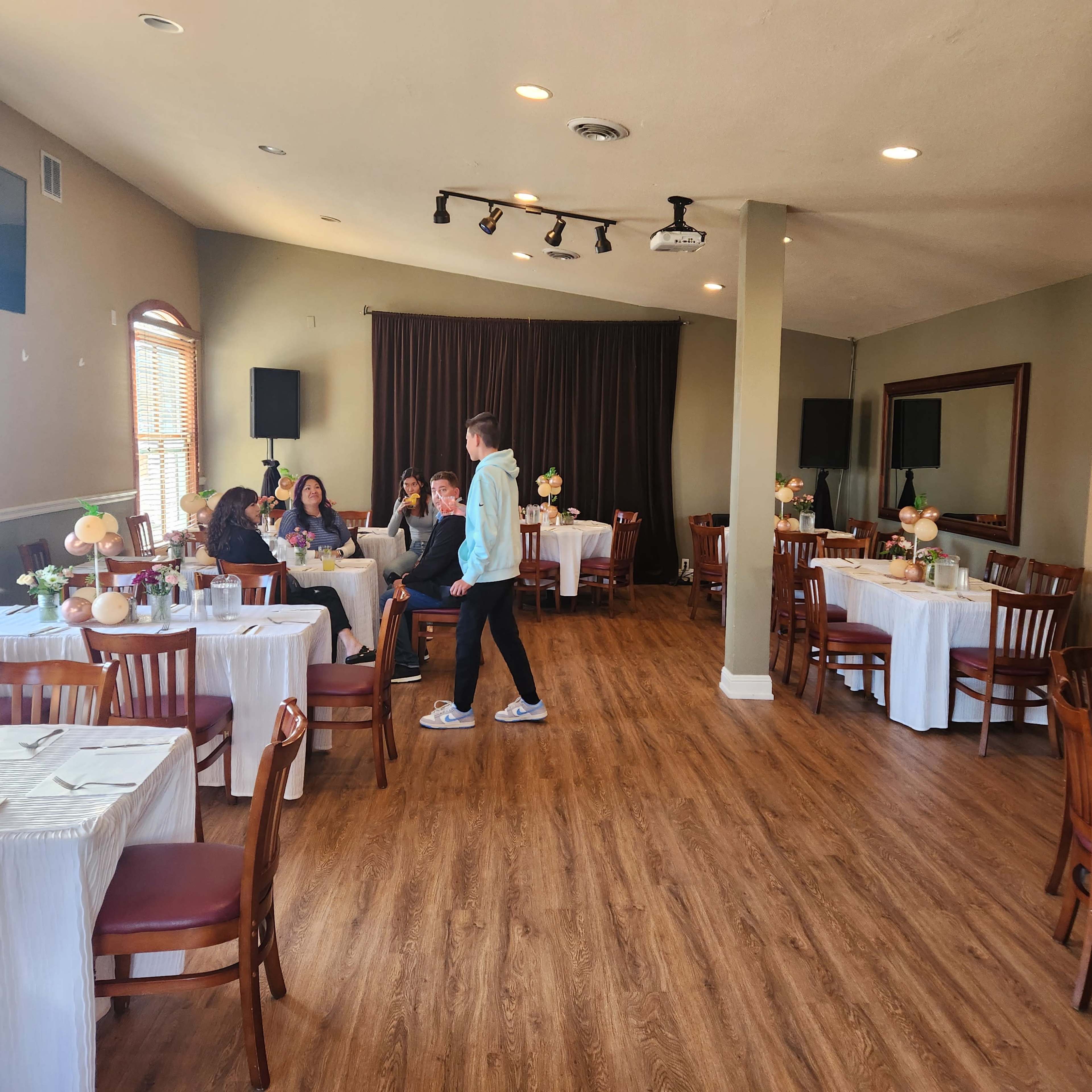 A spacious restaurant interior features several tables set for dining, with a group of patrons at one table and a server attending to them.