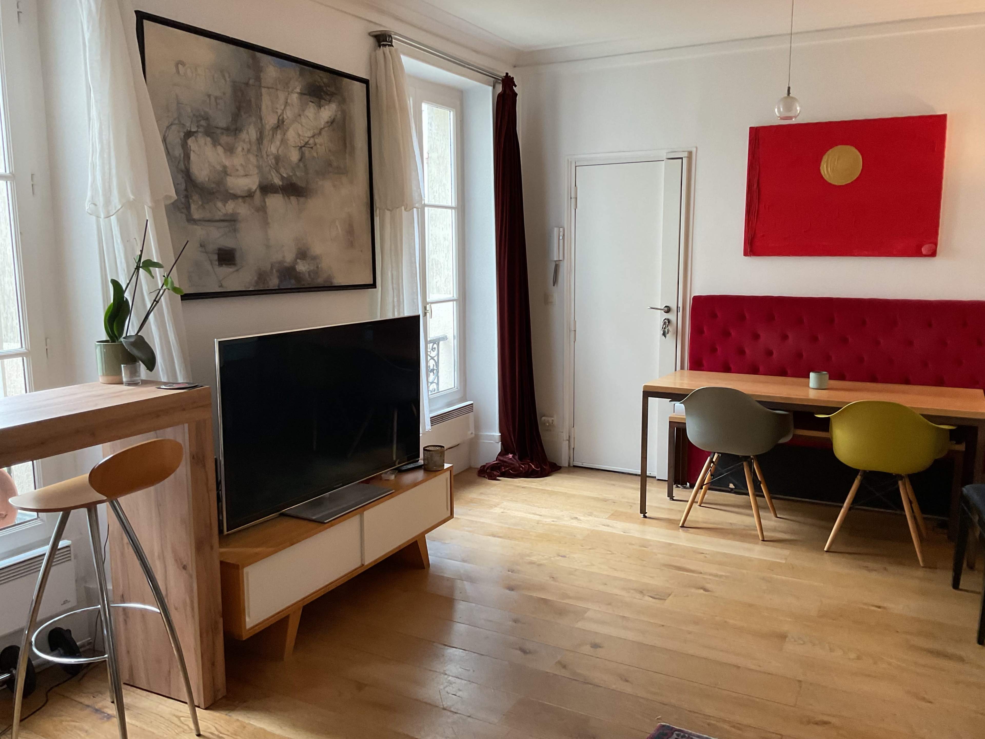 The image shows a bright living space with a wooden floor, a large TV on a modern console, a dining table with colorful chairs, and an abstract painting on the wall.