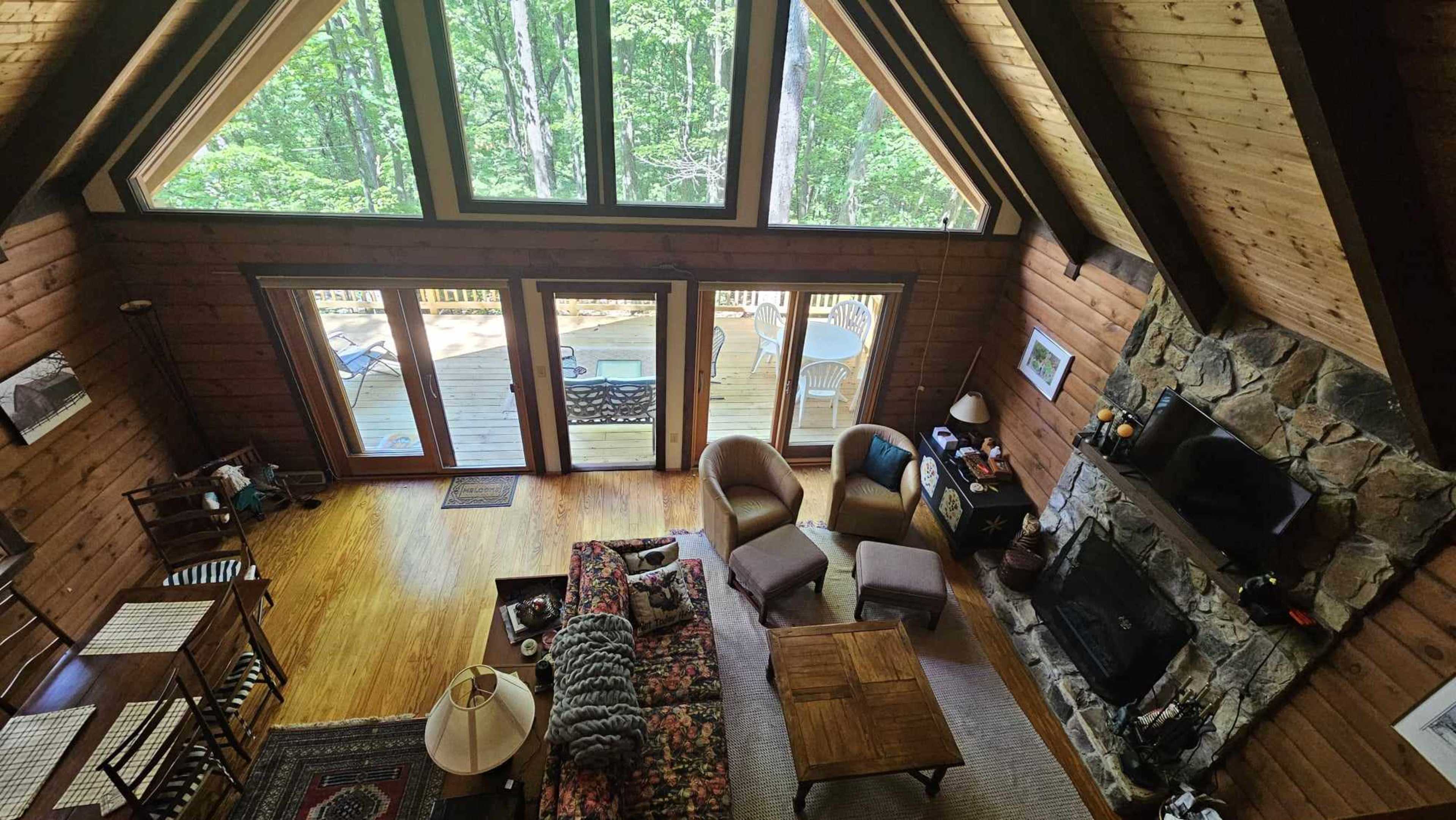 The image shows a cozy, wooden cabin interior with large windows, a stone fireplace, and a spacious seating area overlooking a deck and wooded landscape outside.