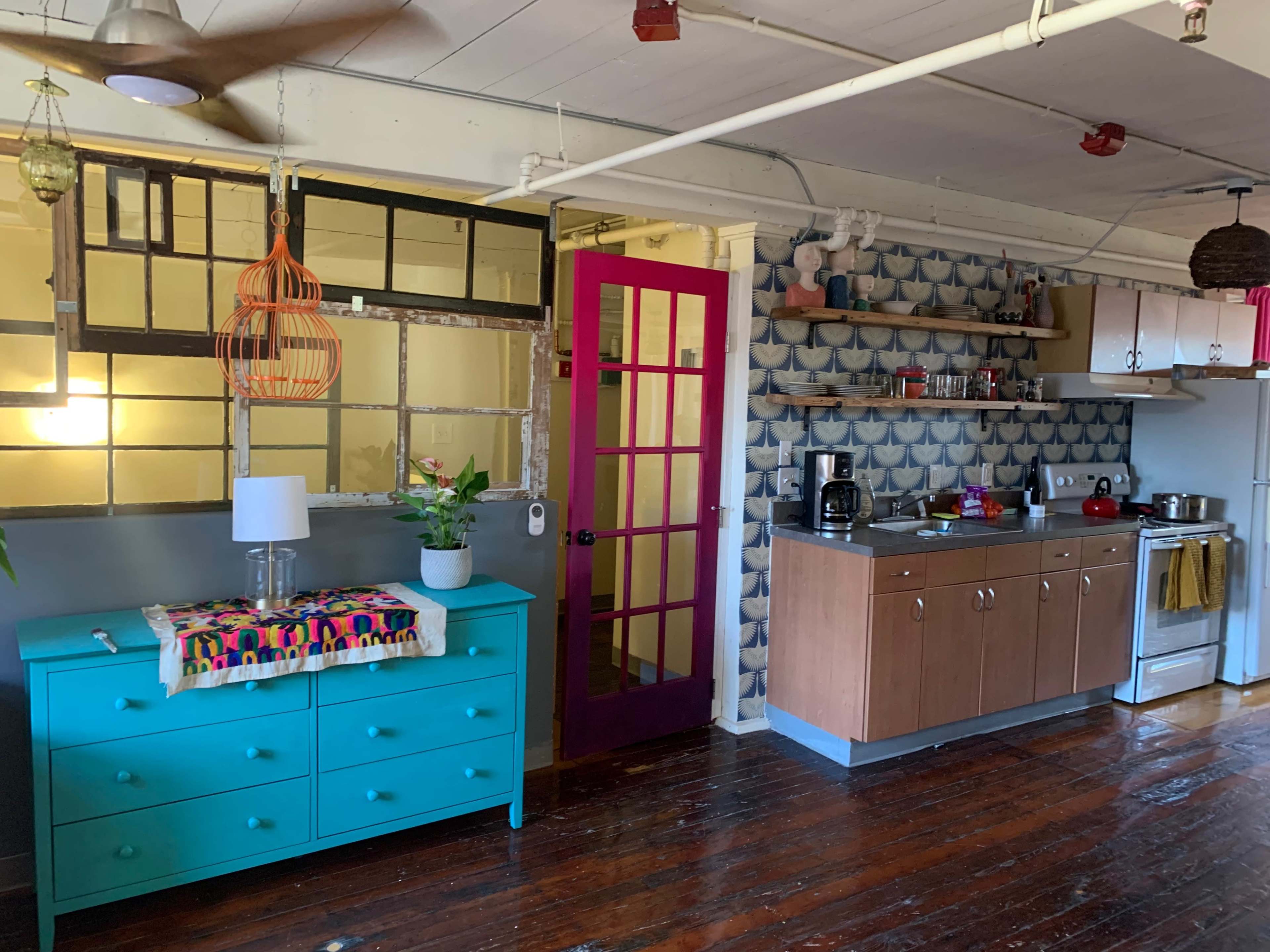 A colorful kitchen and dining area featuring a turquoise dresser, a pink door, and patterned wallpaper with a stove and countertop.
