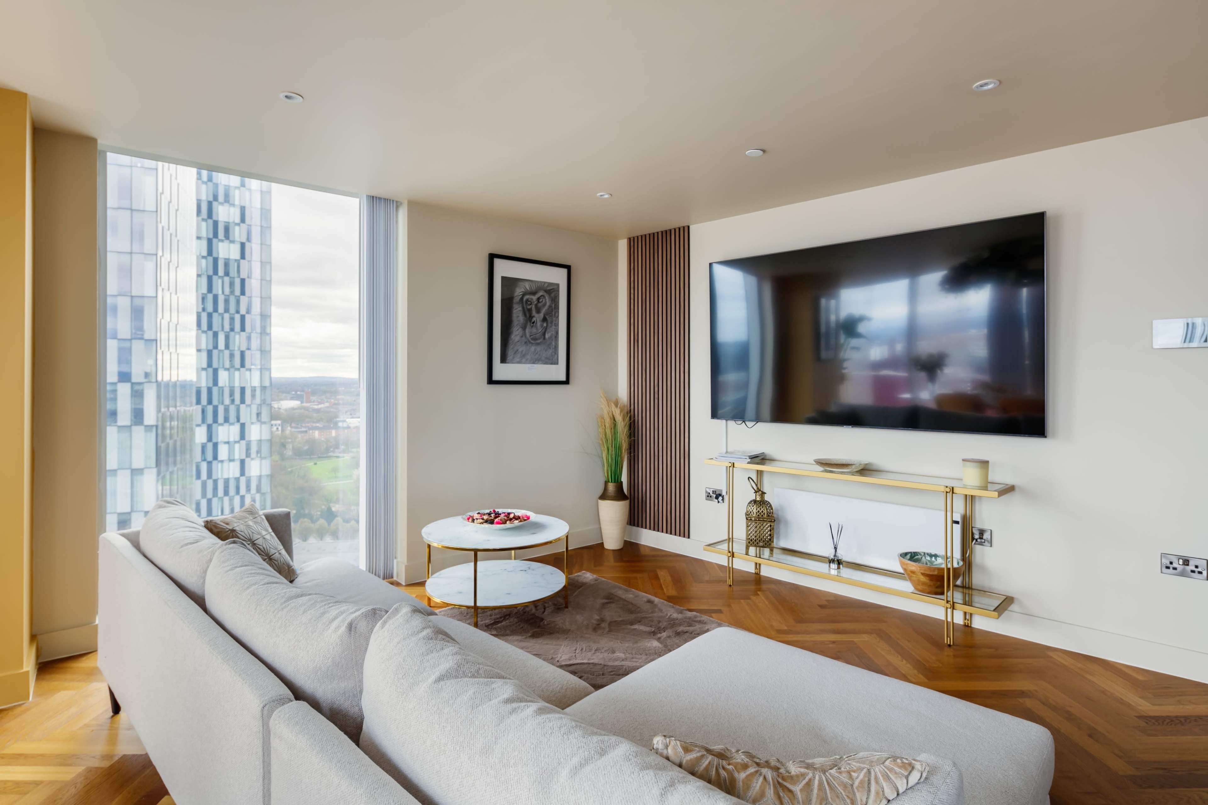 A modern living room features a large television mounted on the wall, a sectional sofa, and a round coffee table, with large windows offering a view outside.