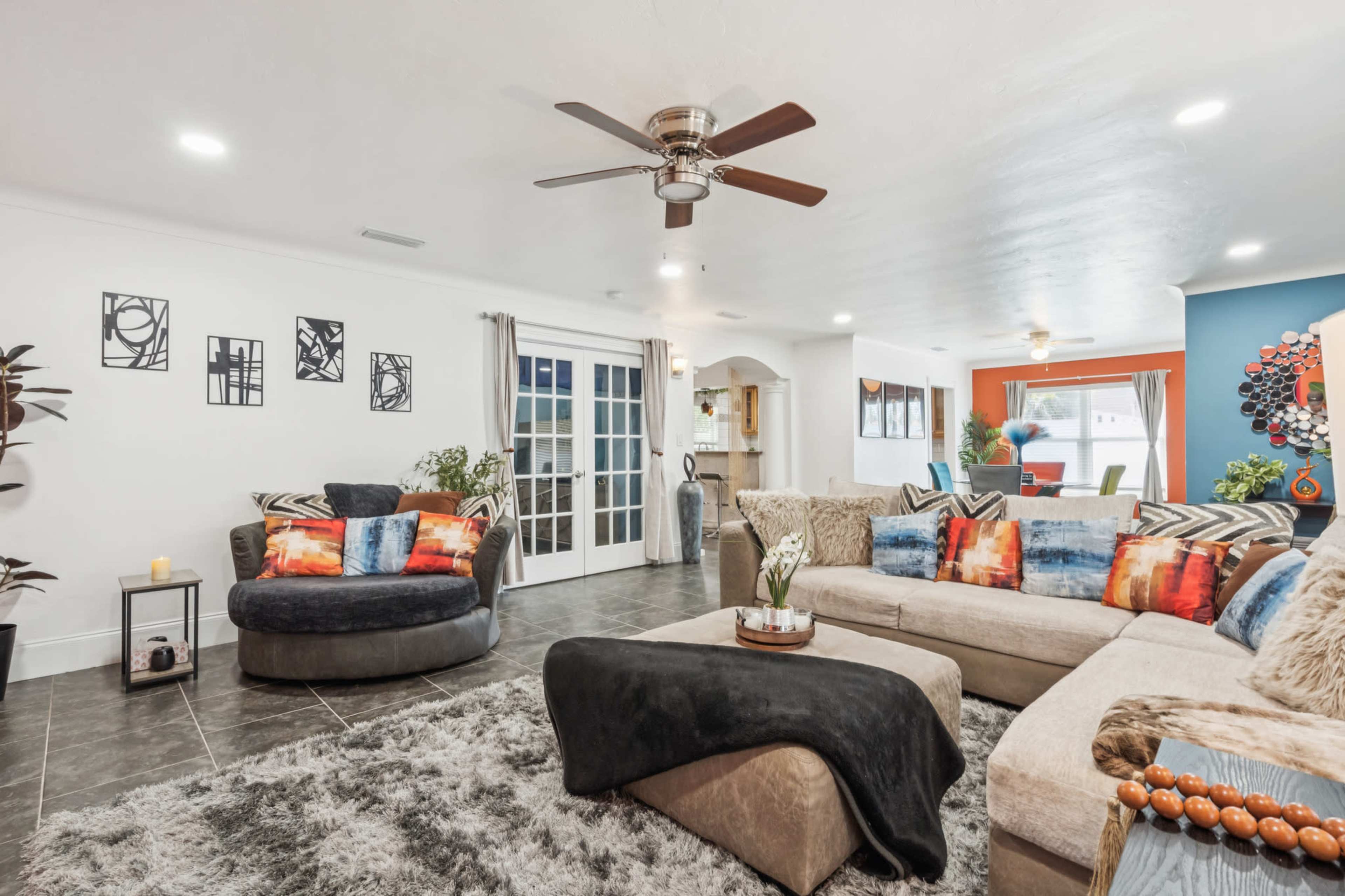 The living room features a large sectional sofa, a circular chair, and a ceiling fan, with a mix of colorful cushions and artwork on the walls.