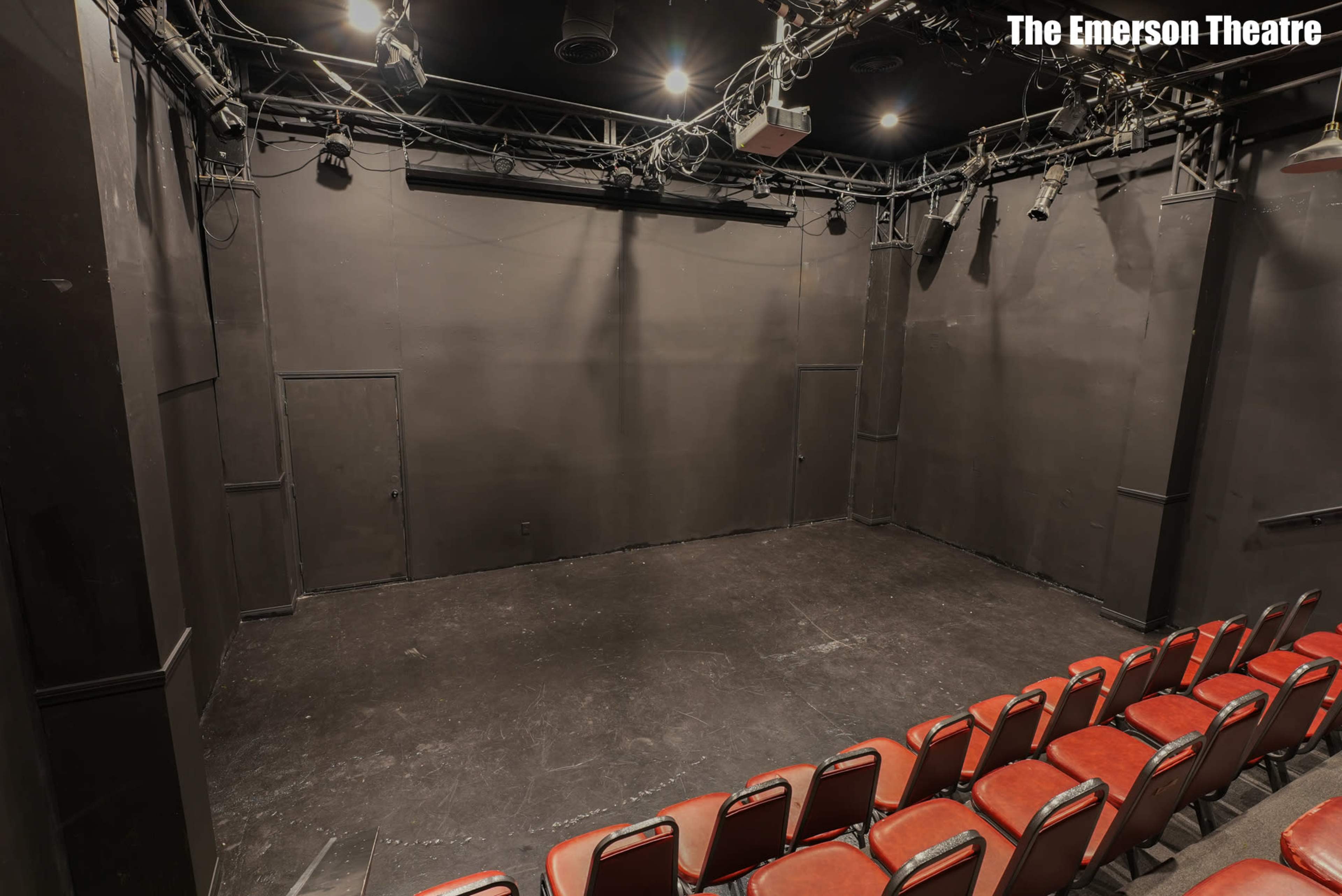 The image shows an empty black box theatre with a seating area in front and stage lighting overhead.