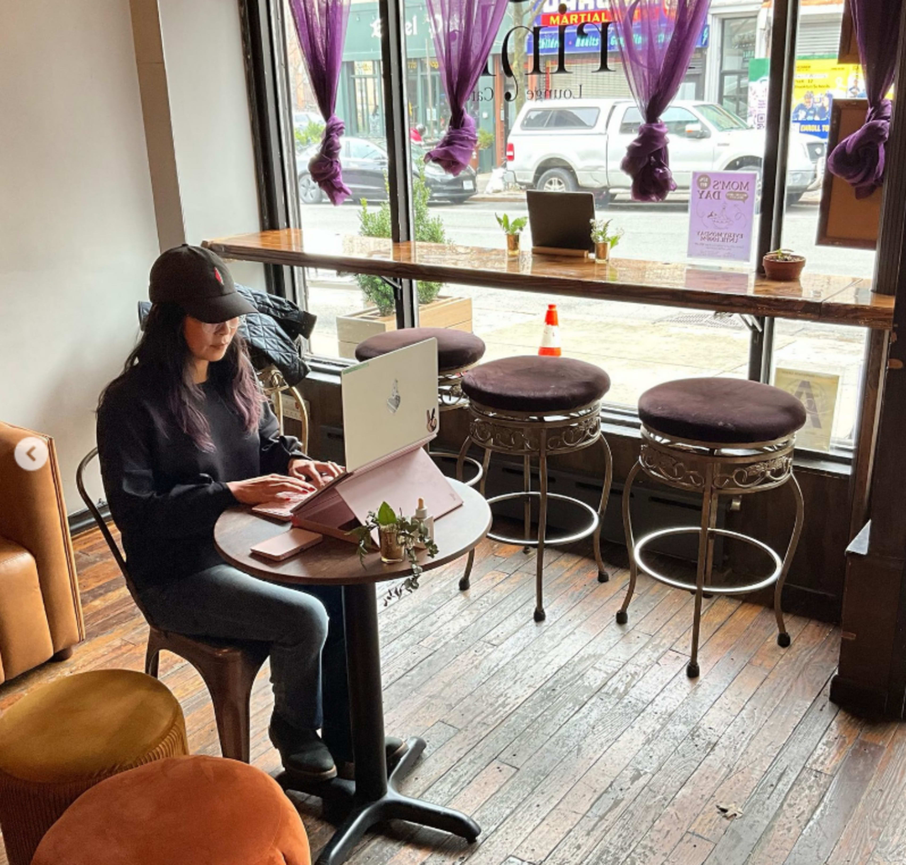 A woman sits at a small table in a café, working on her laptop in front of a large window adorned with purple curtains.