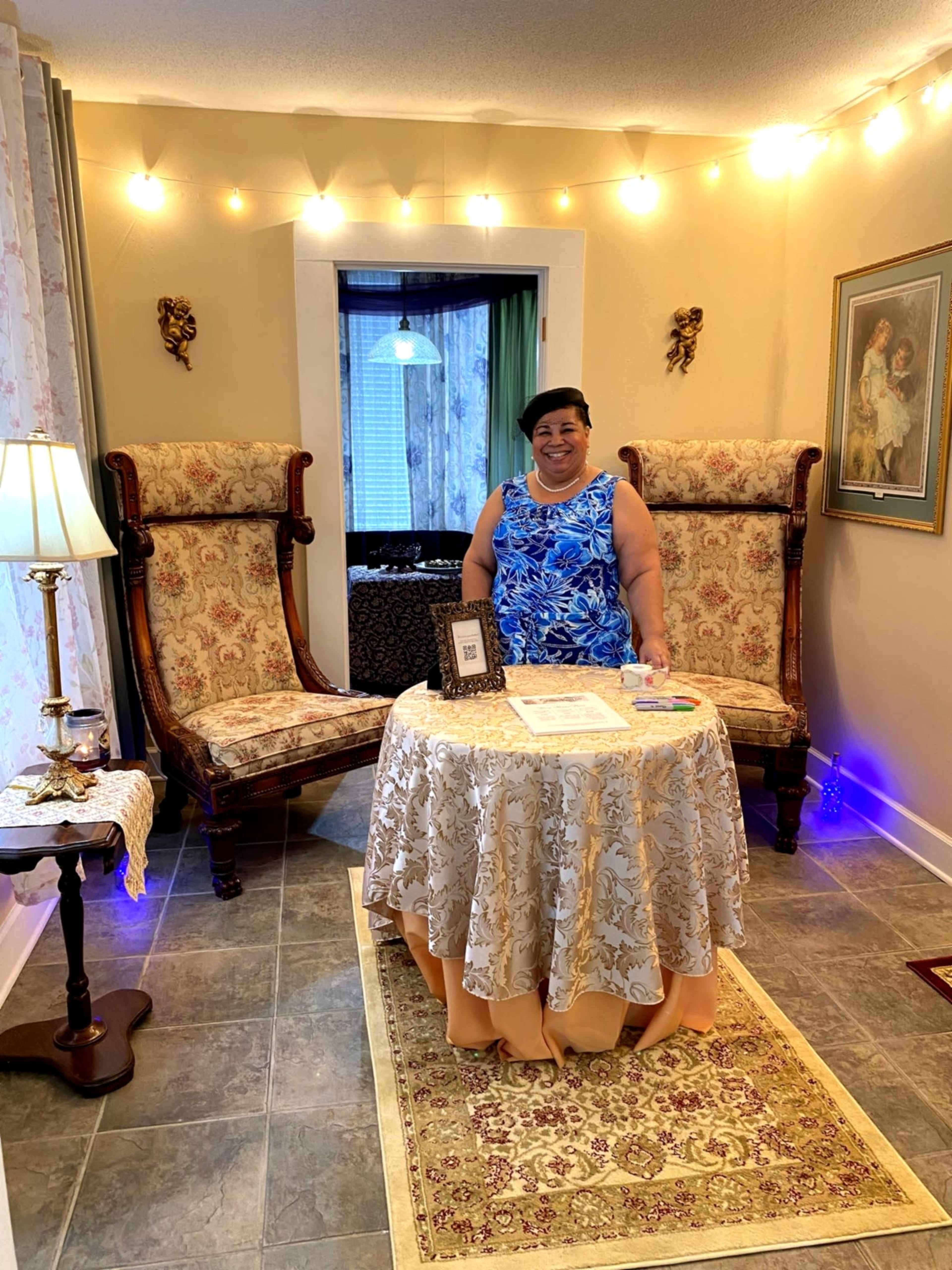 A woman stands smiling behind a round table decorated with a tablecloth in a well-lit room featuring two vintage chairs and soft lighting.