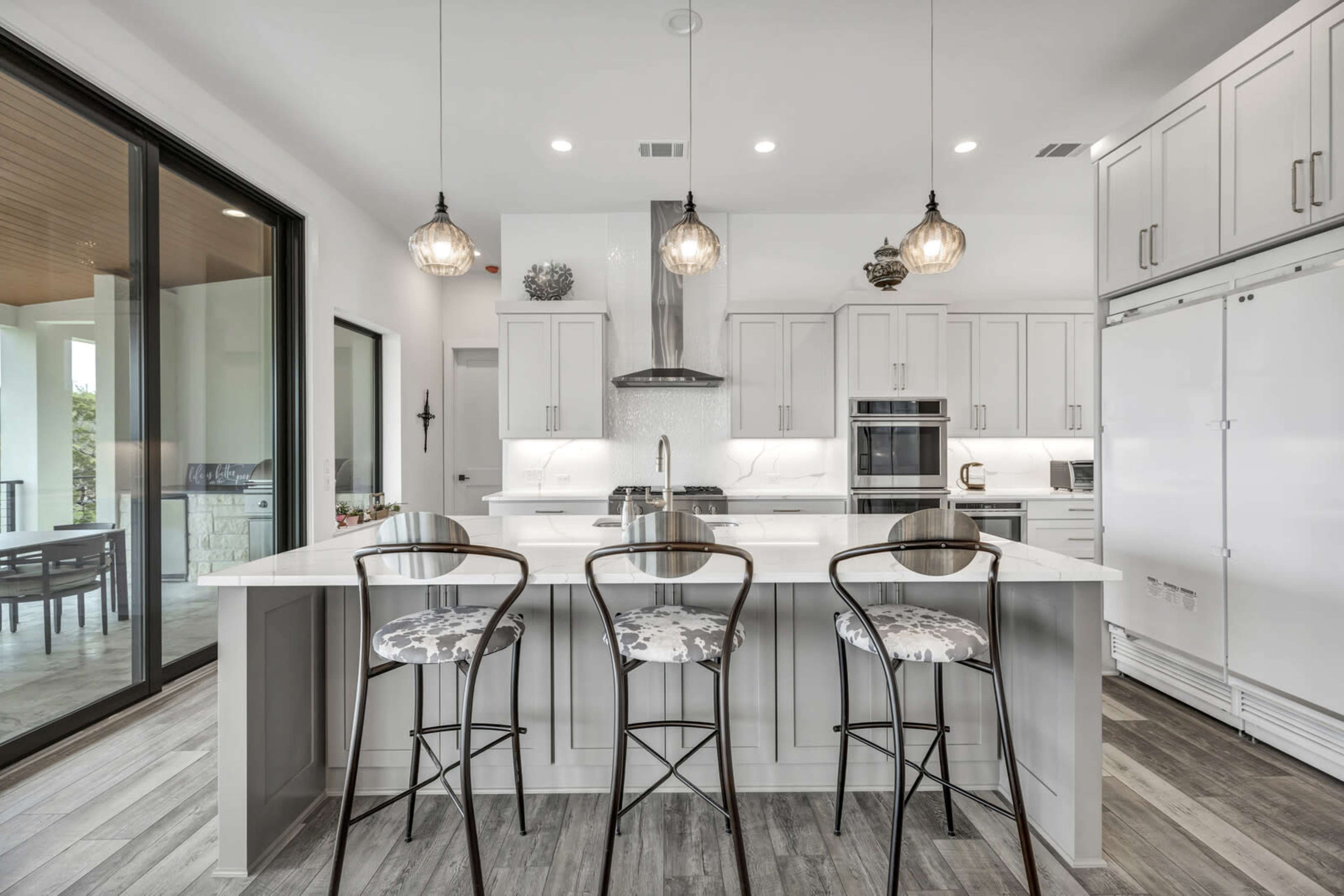 A modern kitchen features white cabinetry, a large island with seating for three, and large windows that provide natural light.