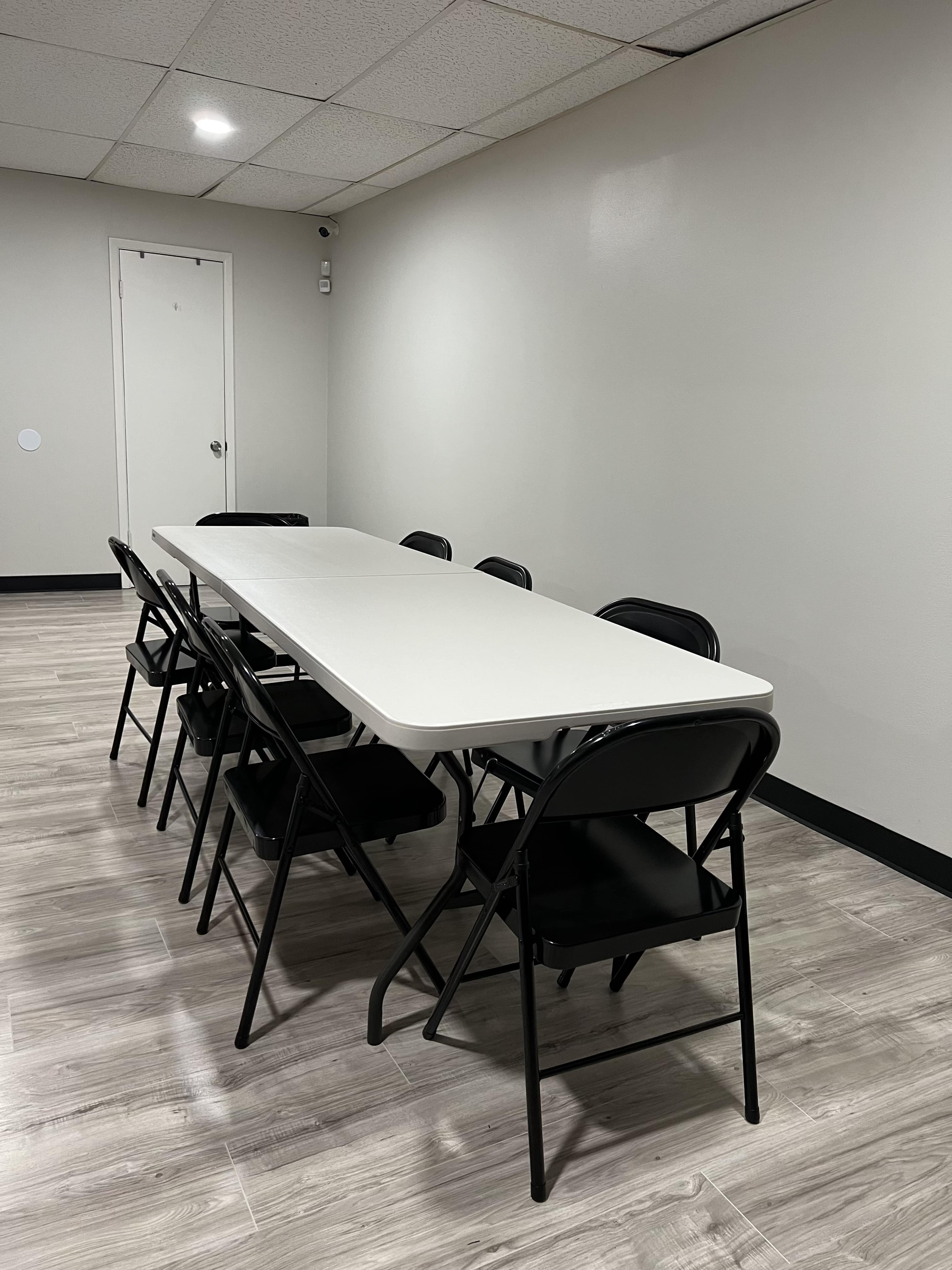 A long, rectangular white table is set in a brightly lit room with six black folding chairs arranged around it.