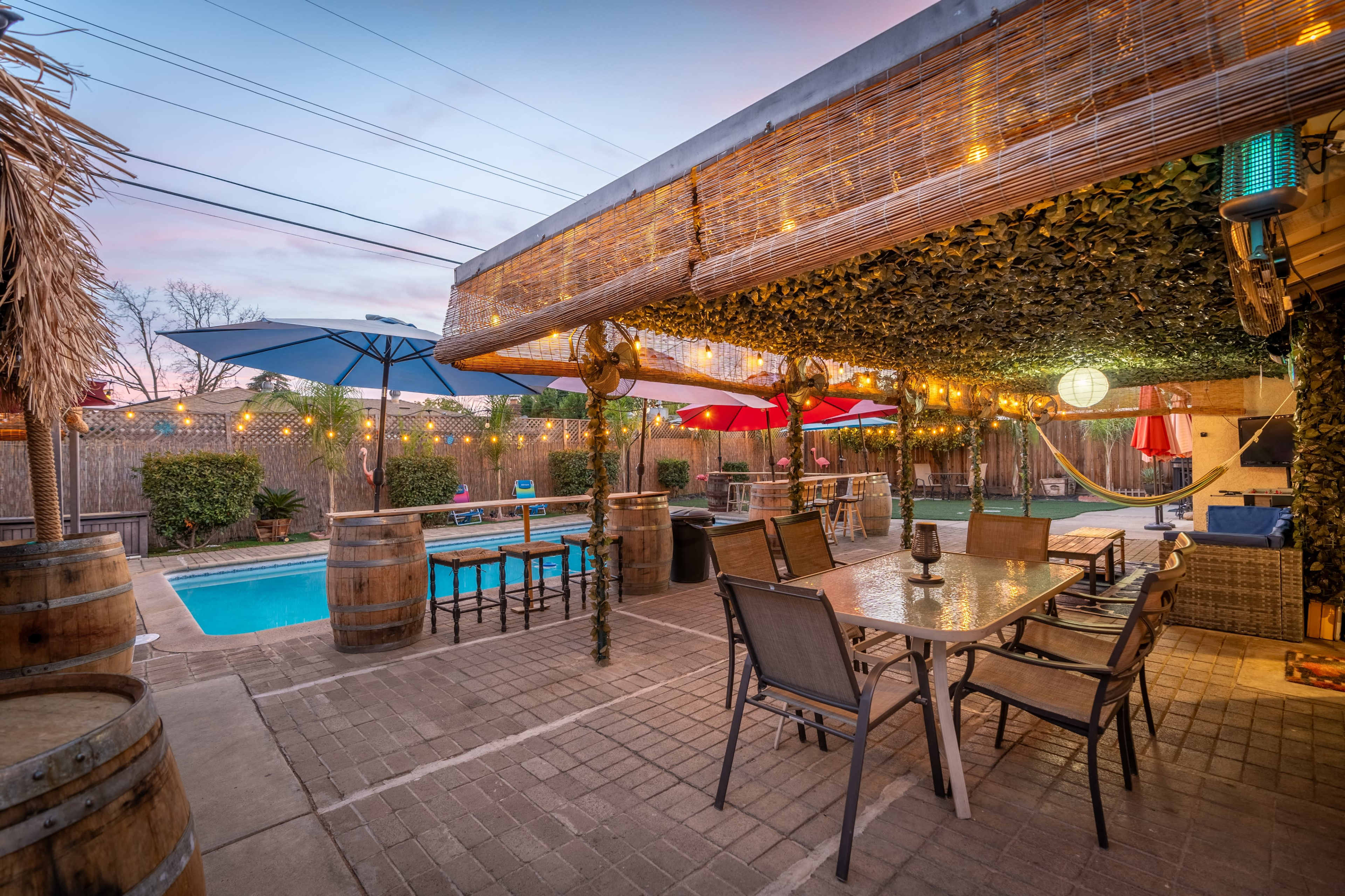 The outdoor space features a pool surrounded by seating areas, a dining table with chairs, and shaded umbrellas, all under decorative string lights.