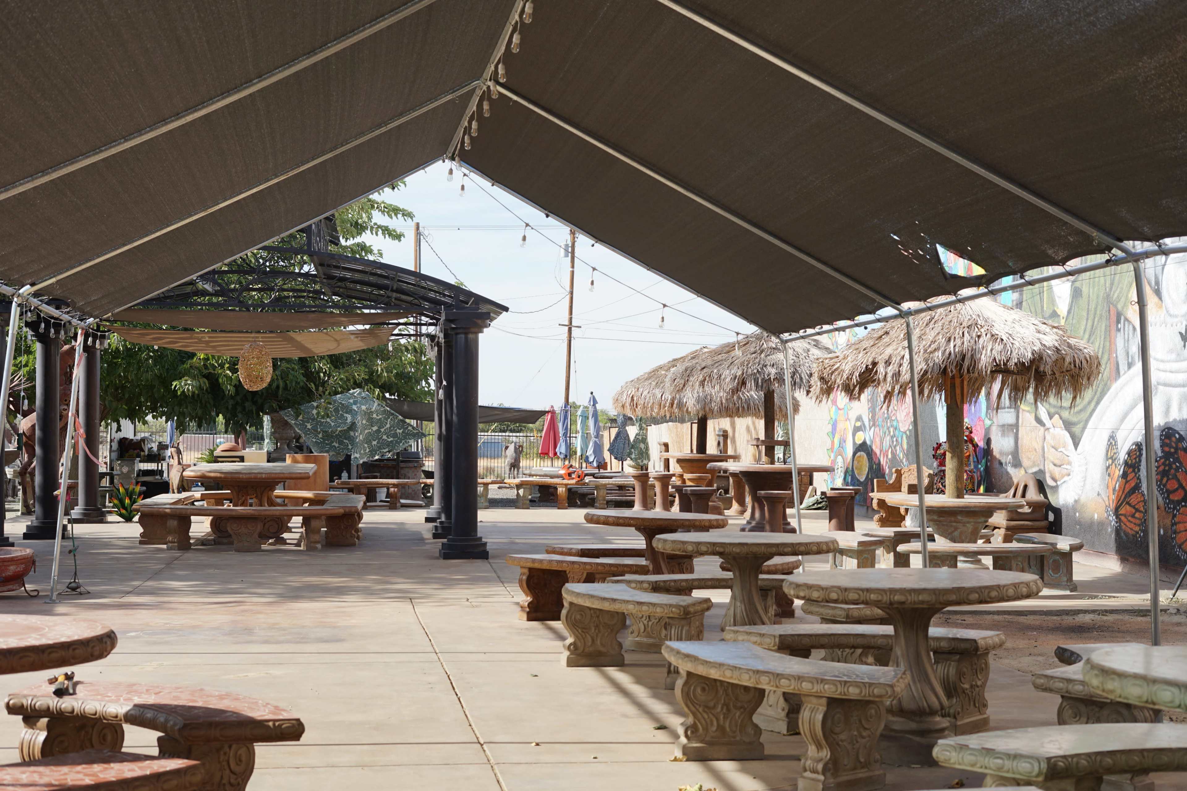 The image shows an outdoor seating area with stone tables and a shaded canopy, featuring decorative murals on the walls and straw-covered tables.