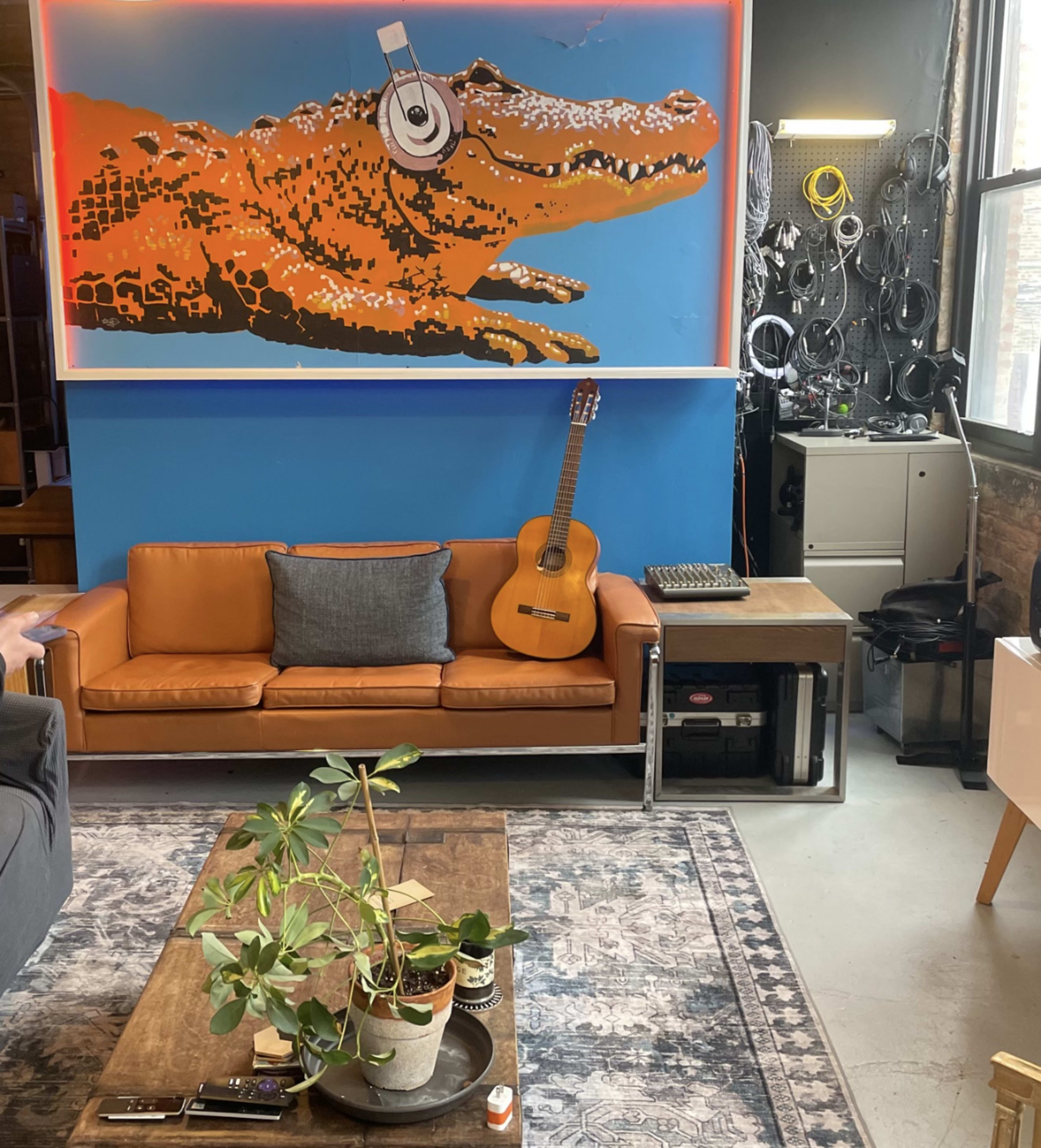The image shows a cozy living space with a brown leather couch, an acoustic guitar hanging on the wall, a wooden coffee table with a plant on it, and a large colorful alligator mural.