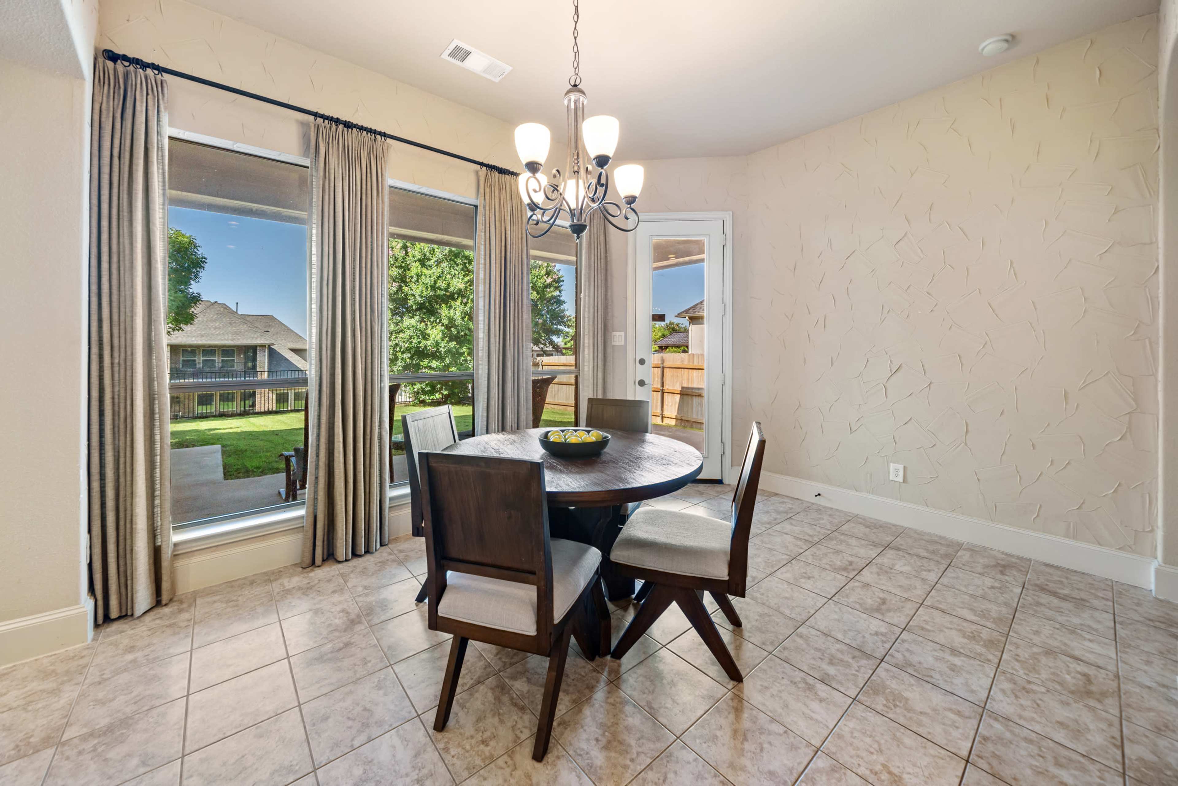 A dining area features a round table with four chairs, large windows showing a lawn outside, and a chandelier hanging from the ceiling.