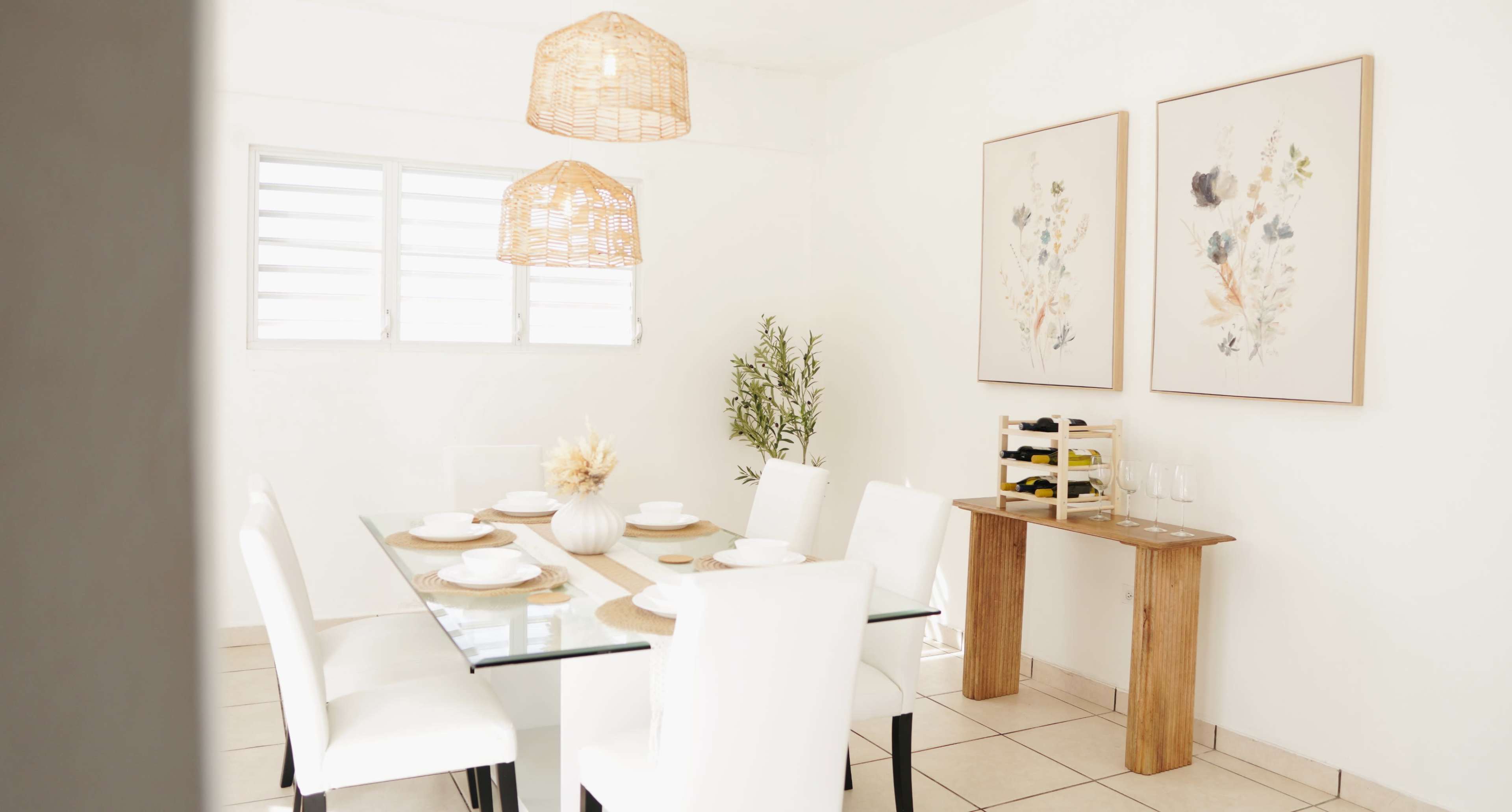 A modern dining room features a glass table with white chairs, pendant lights, and floral artwork on the walls.
