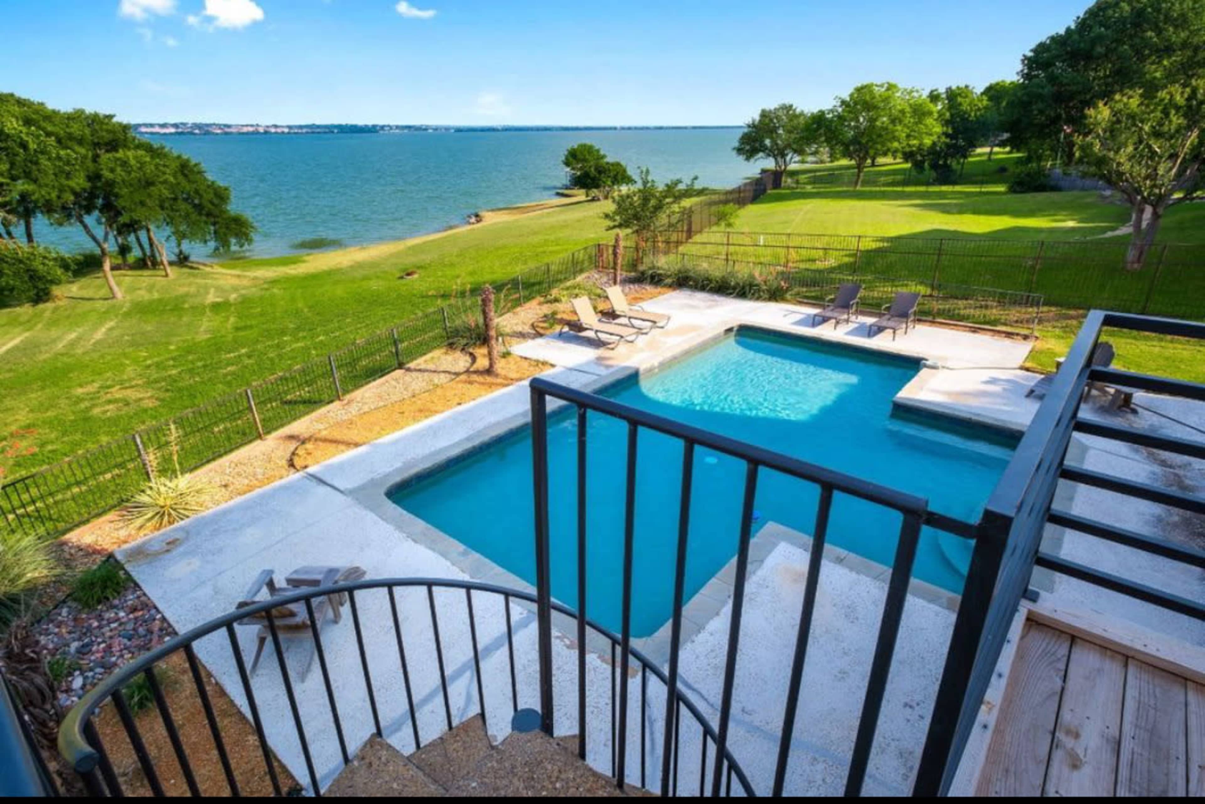 The image shows a backyard with a swimming pool overlooking a large body of water, surrounded by green grass and trees.