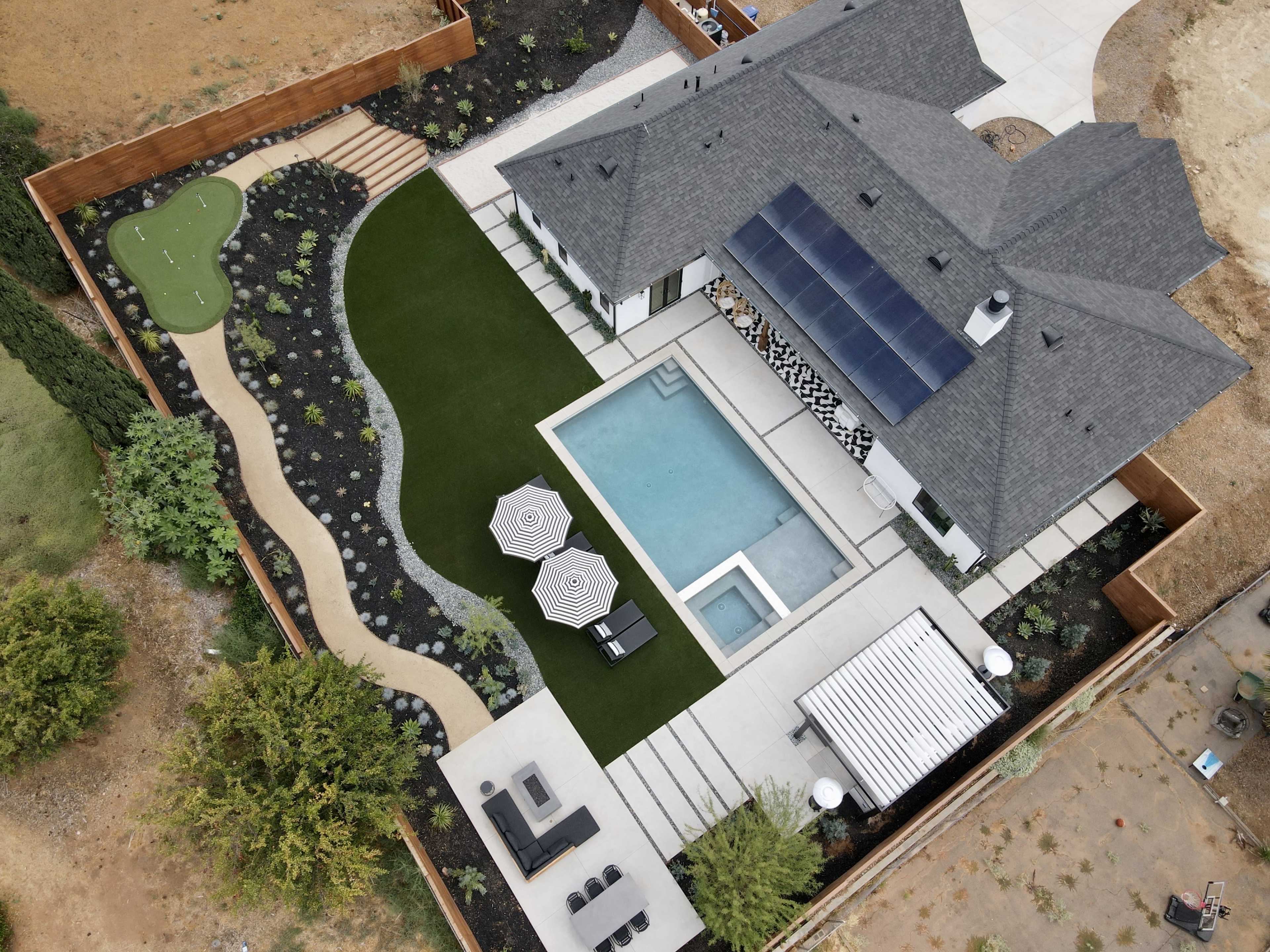 An aerial view of a contemporary house featuring a swimming pool, landscaped lawn, and outdoor seating areas surrounded by a wooden fence.