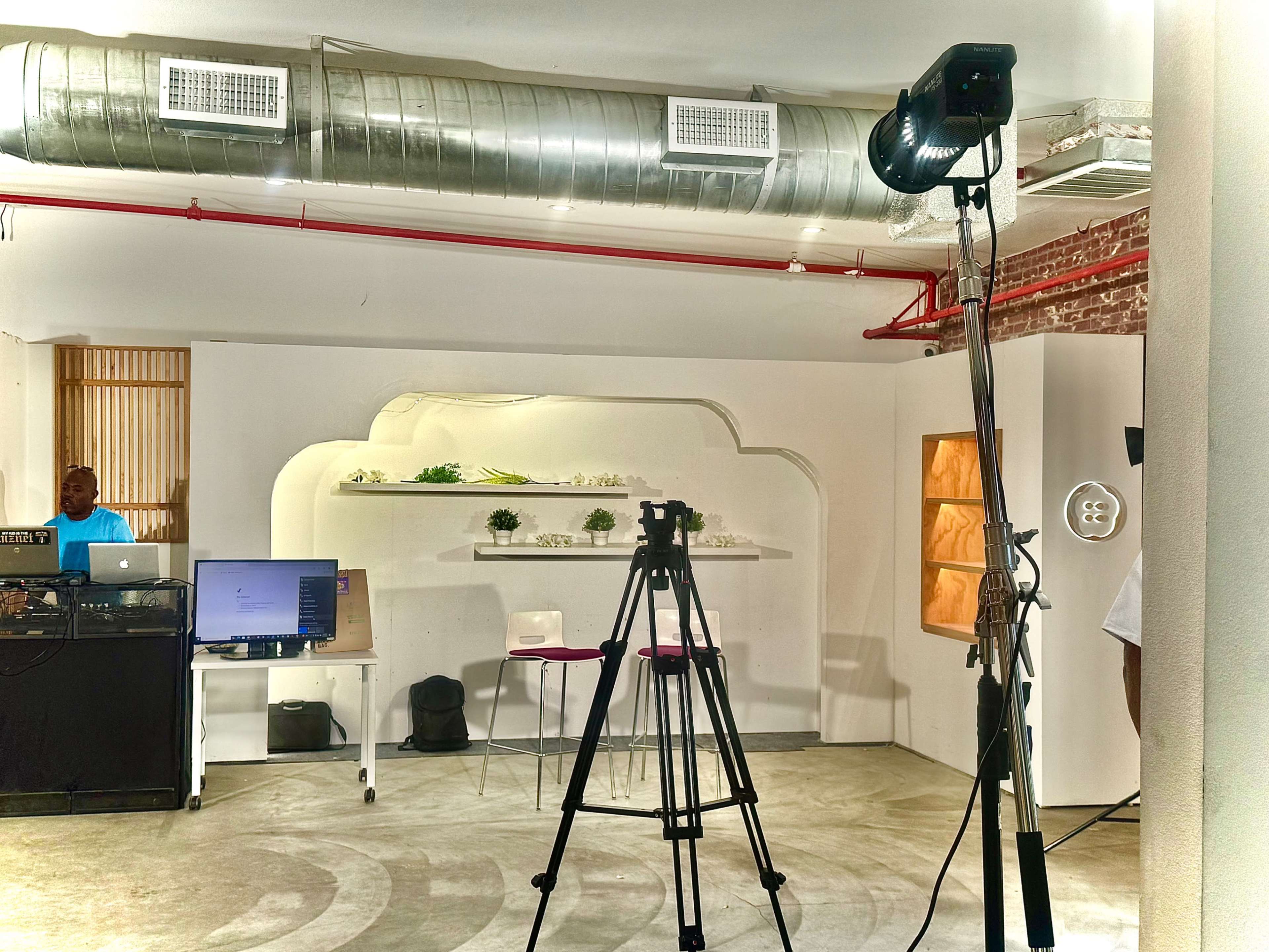 The image shows a modern indoor space with a tripod set up in the foreground, a desk with a computer on the left, and minimalistic decorative shelves in the background.