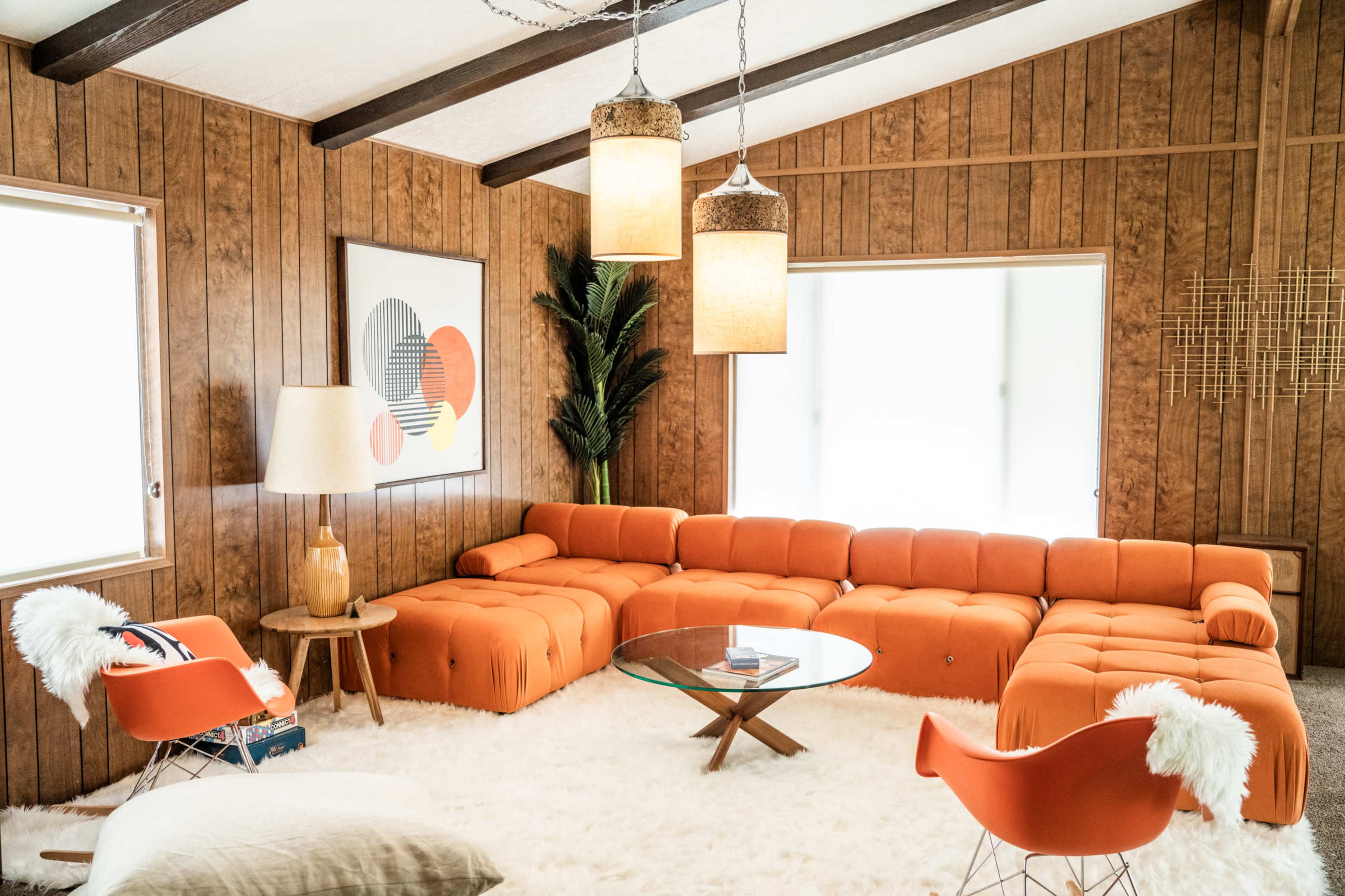 The image shows a modern living room with an orange sectional sofa, a glass coffee table, and wooden paneling on the walls.
