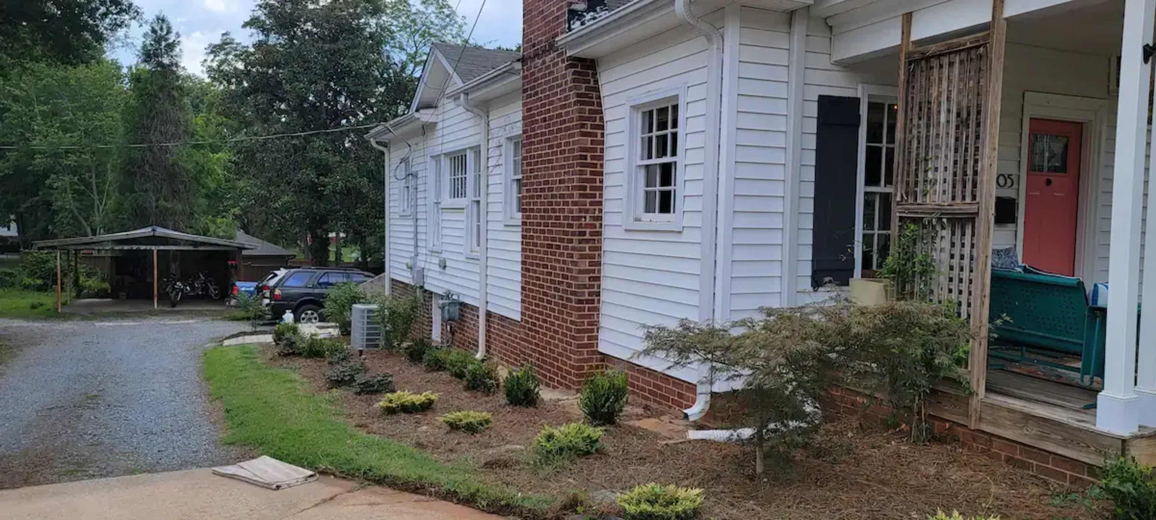 A white house with a brick chimney, a gravel driveway leading to a carport, and a garden area with shrubs and plants along the side.