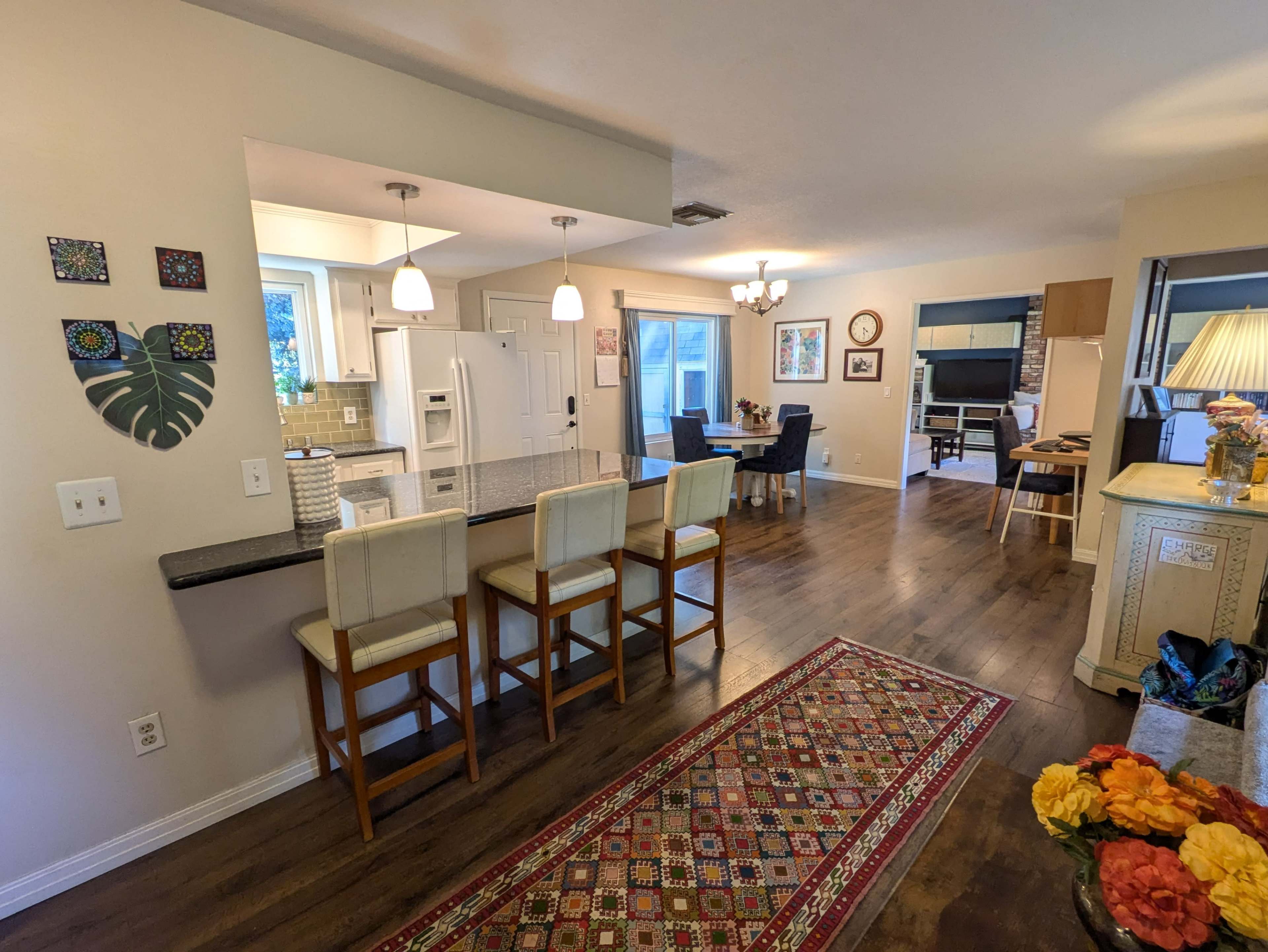 The image shows a bright, open-concept kitchen and living area with bar seating, a dining table, and a decorative rug on the hardwood floor.