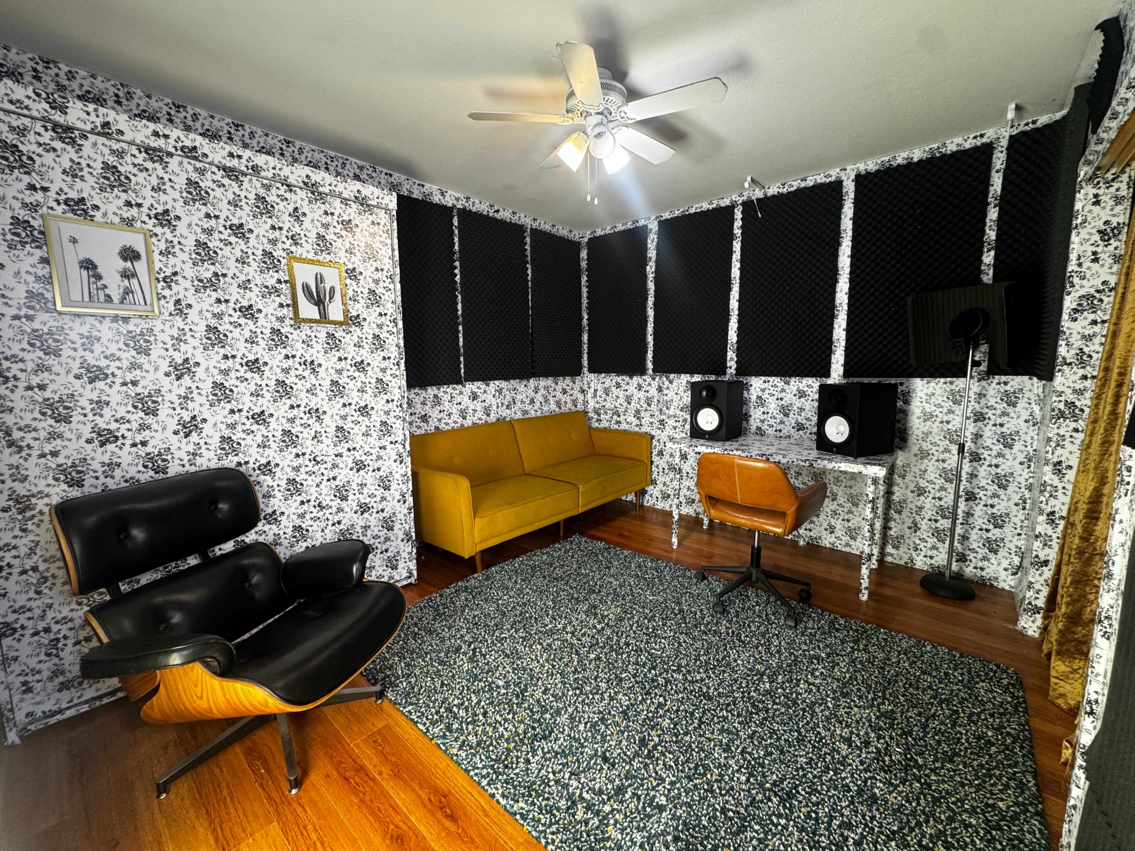 The image depicts a music studio featuring a yellow couch, a black lounge chair, two studio monitors on a desk, and patterned wallpaper.
