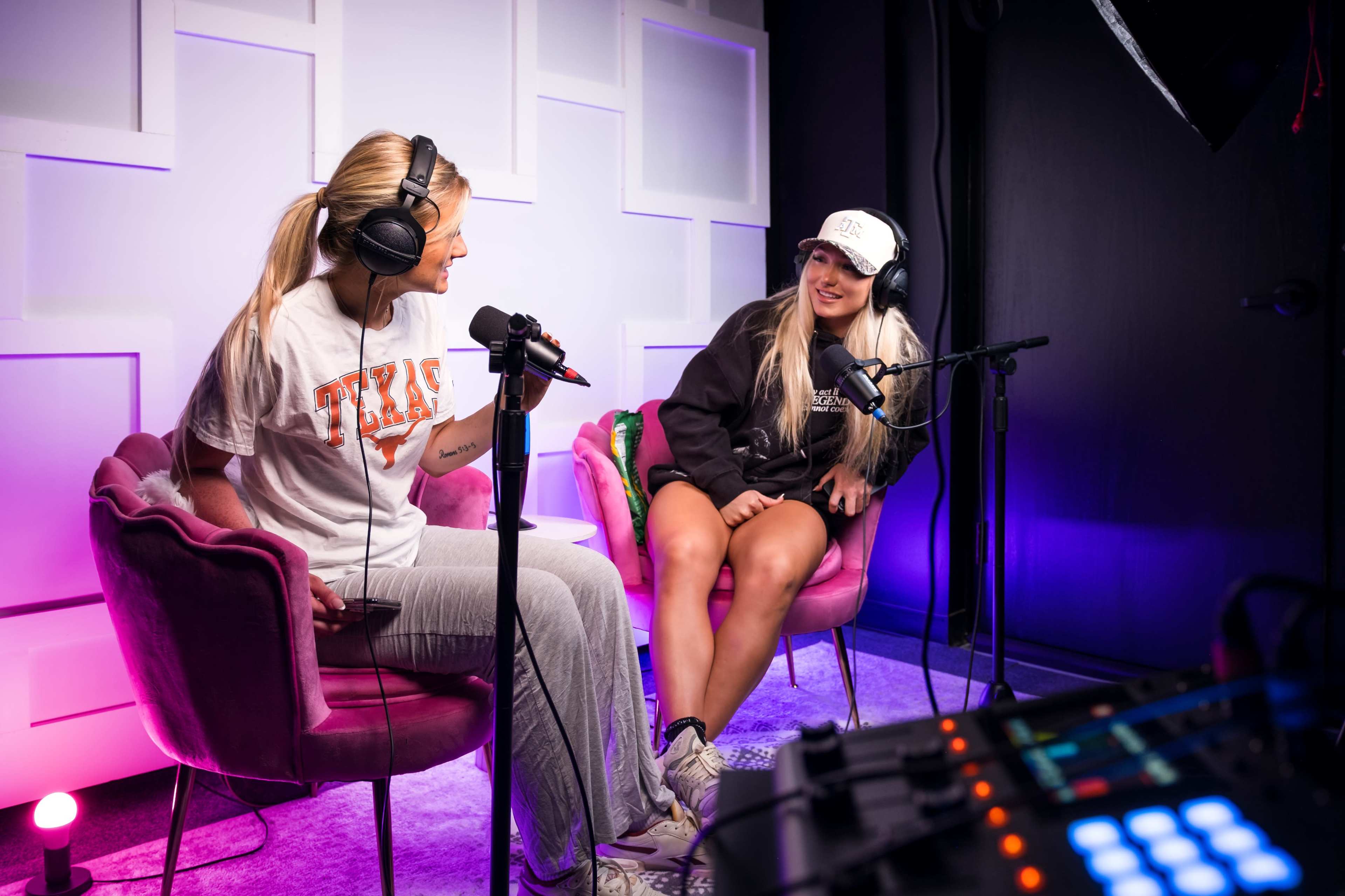 Two women are seated in pink chairs, wearing headphones and microphones, engaged in a conversation in a brightly lit studio.