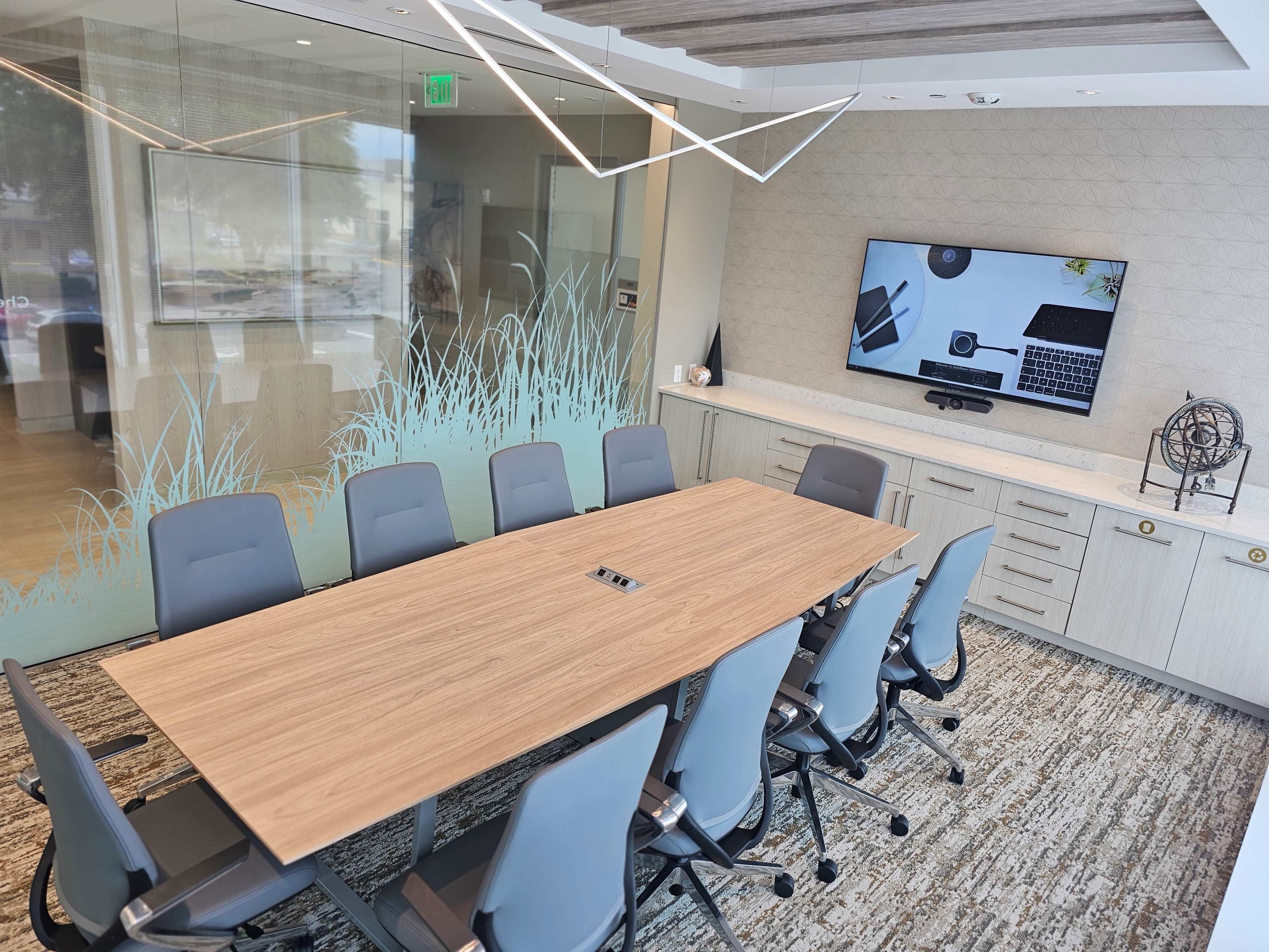 A modern conference room equipped with a large wooden table, ergonomic chairs, and a wall-mounted screen displaying a laptop and office supplies.