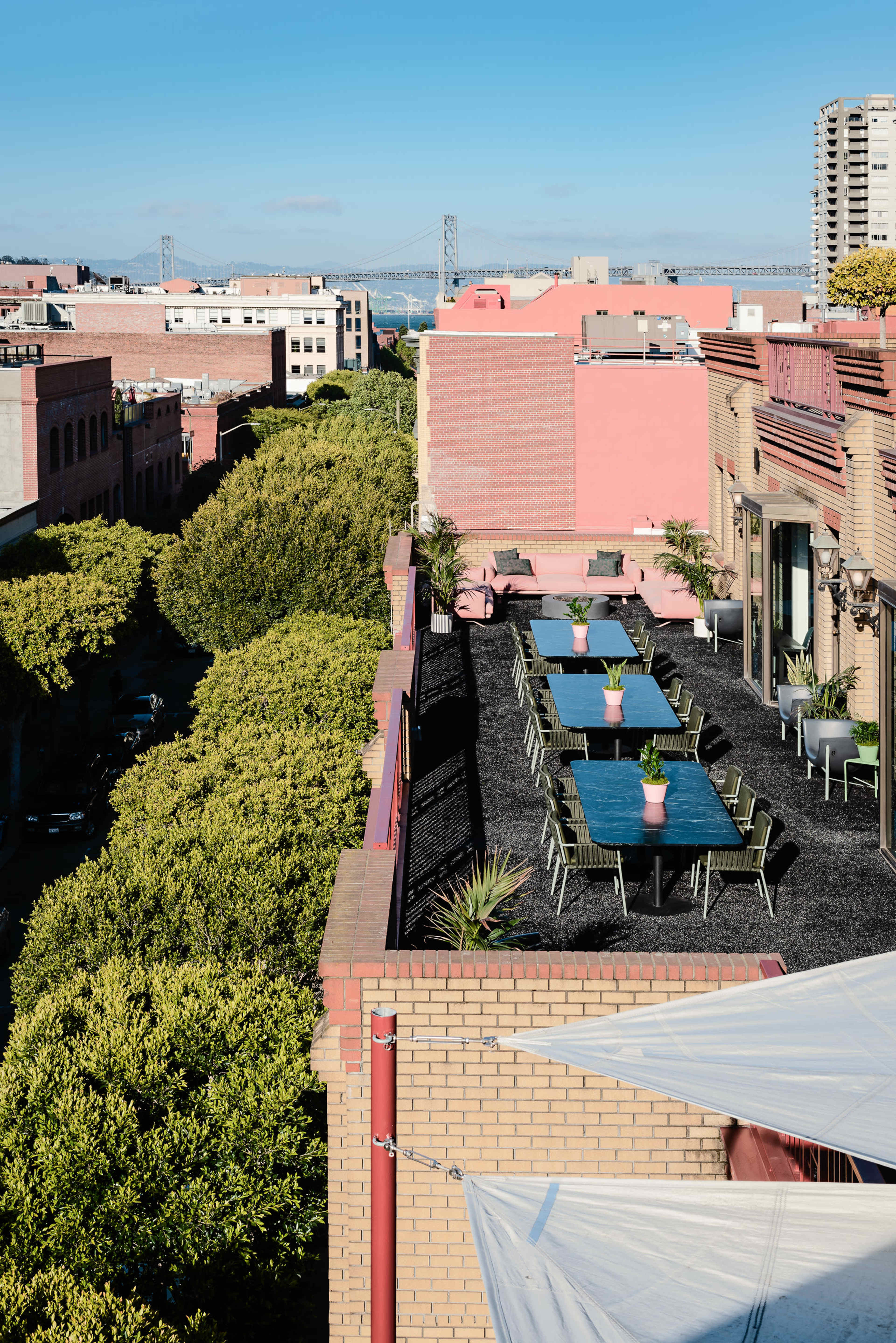The image shows a rooftop terrace with dining tables surrounded by greenery, overlooking a cityscape and a bridge in the background.