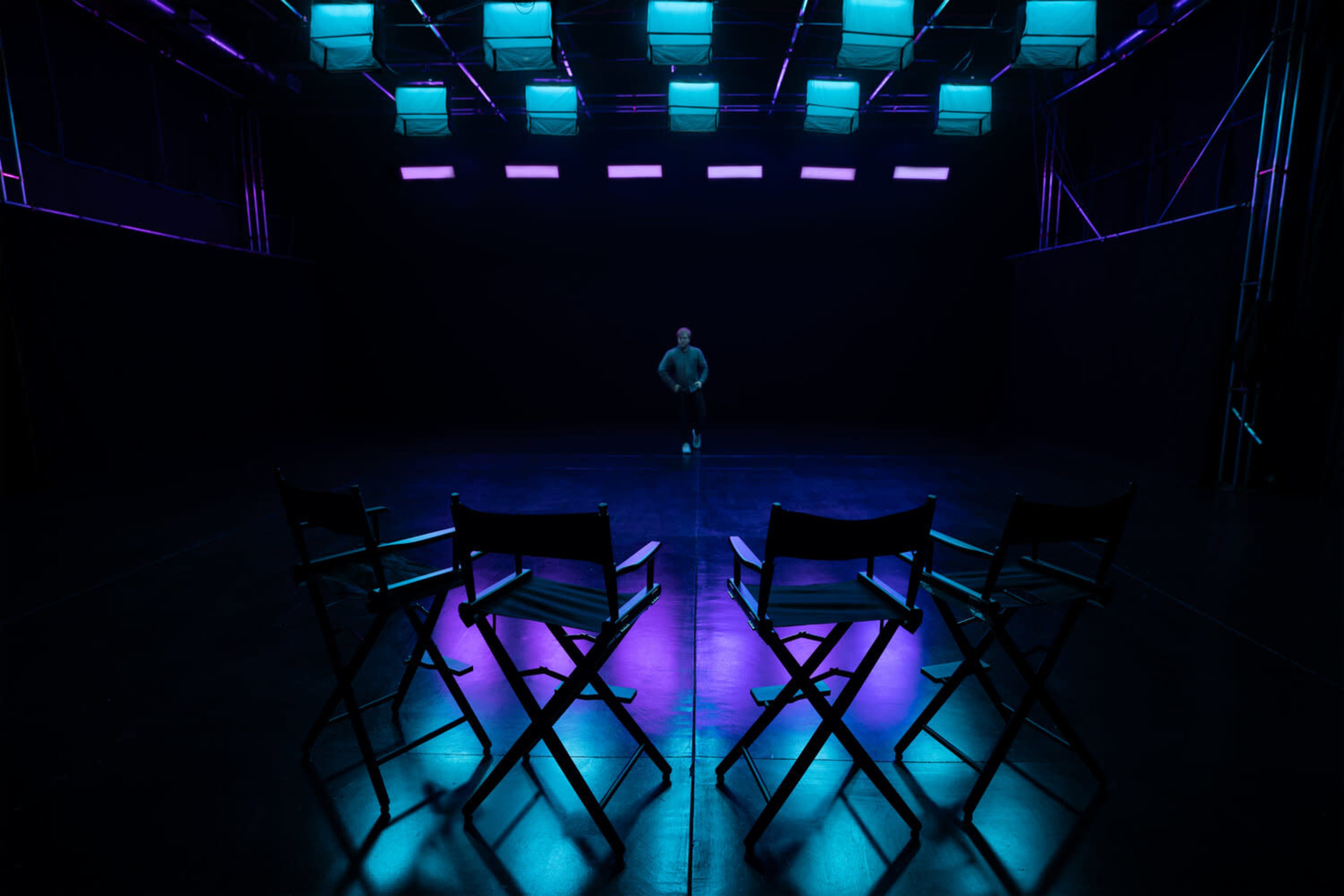 A single figure walks in a dimly lit performance space with four empty director's chairs arranged in the foreground.
