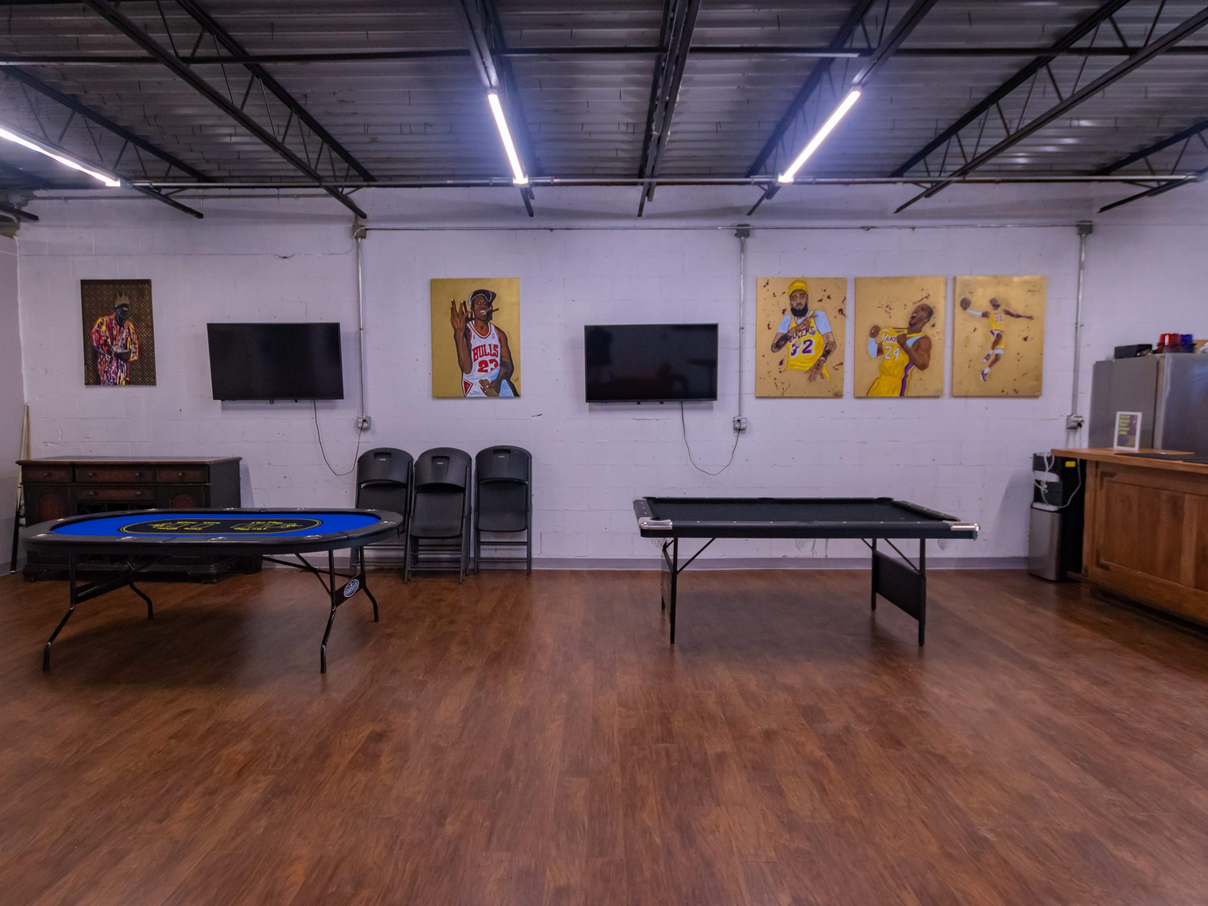 A recreational room with a blue poker table on the left, a black pool table on the right, and two mounted televisions along with framed artwork of basketball players on the walls.