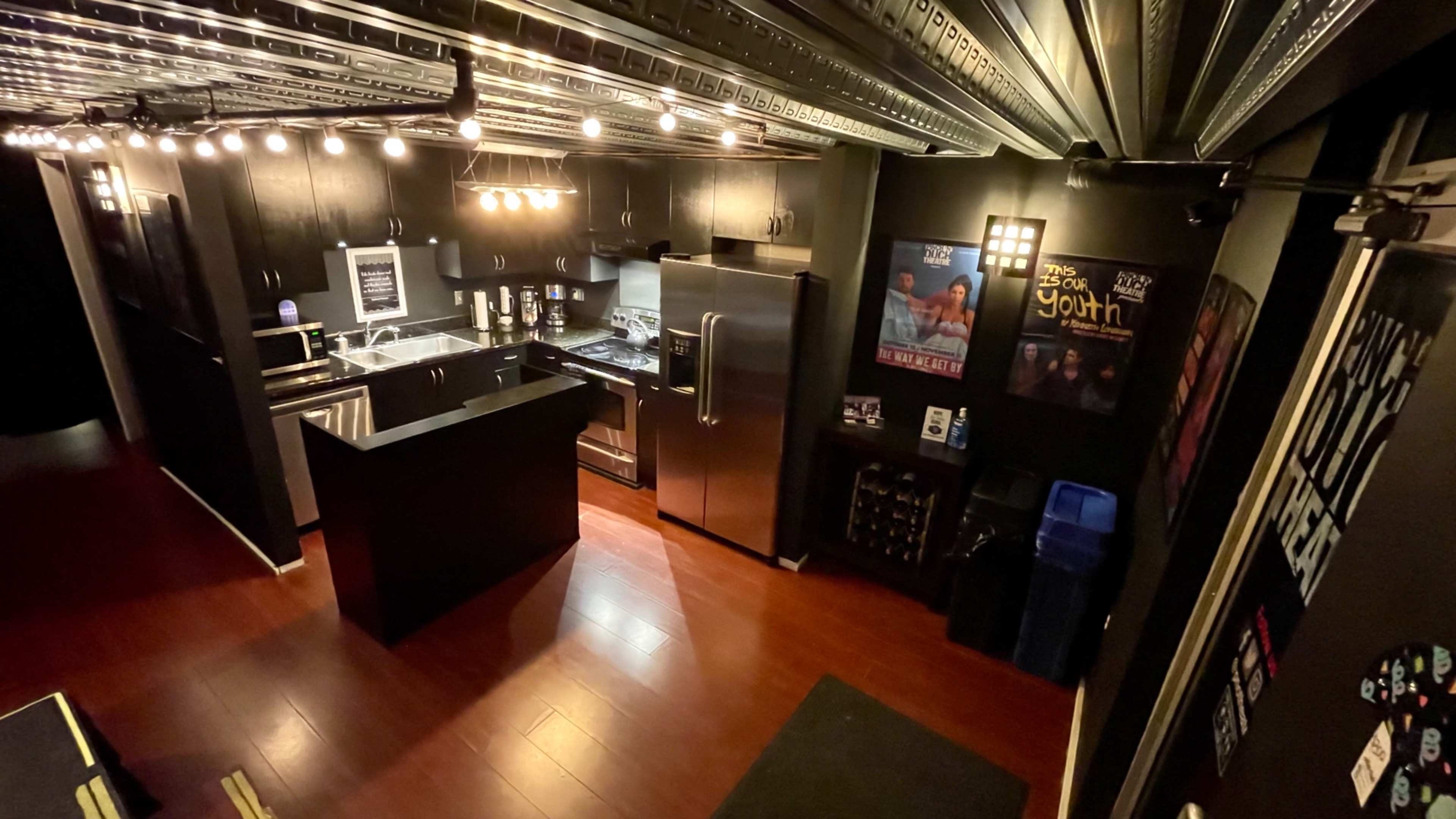 The image shows a modern kitchen with stainless steel appliances, a black island countertop, and dark walls illuminated by overhead lighting.