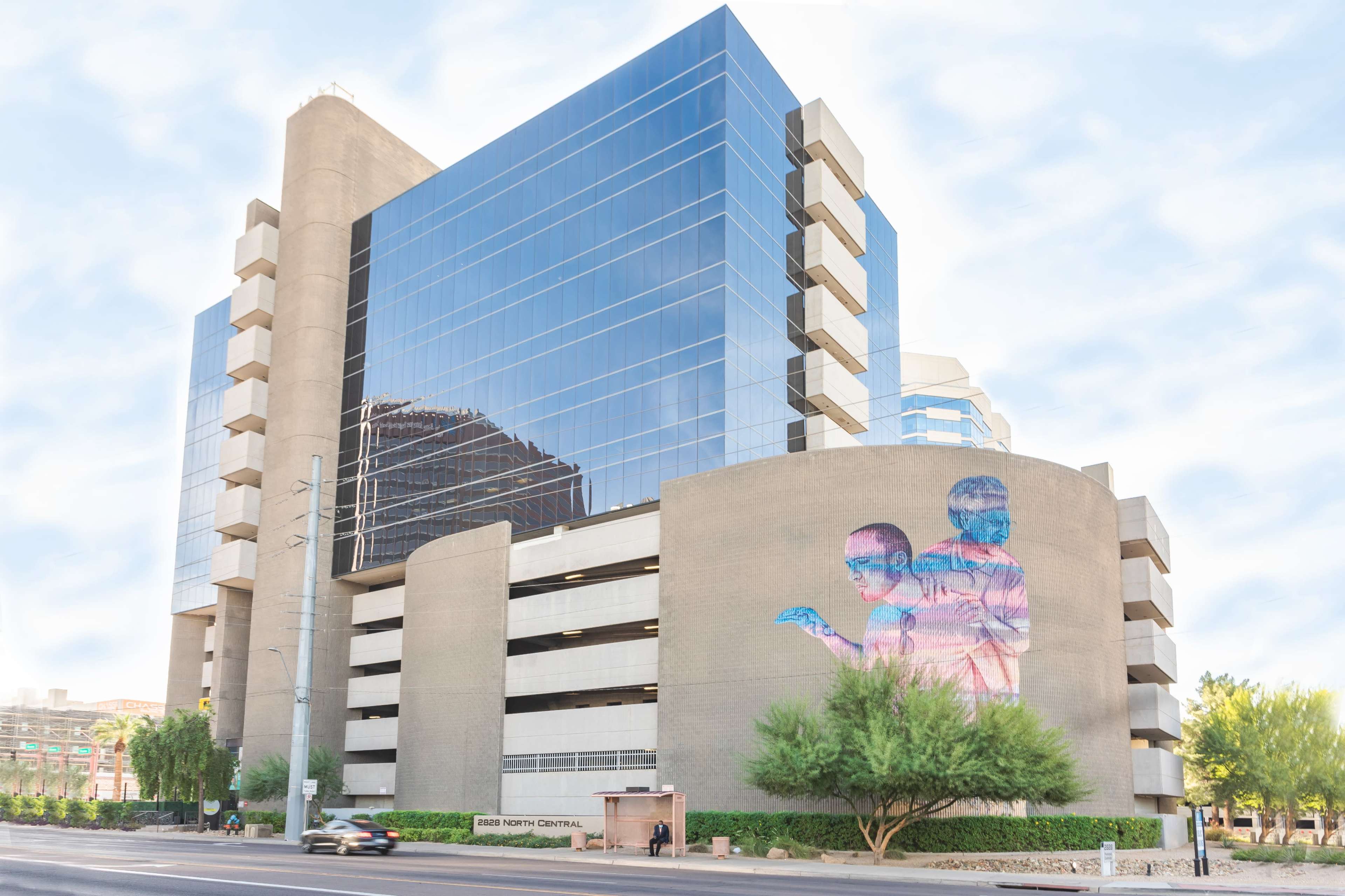 A modern multi-story parking garage features a large mural of two figures on its side, surrounded by greenery and clear blue skies.