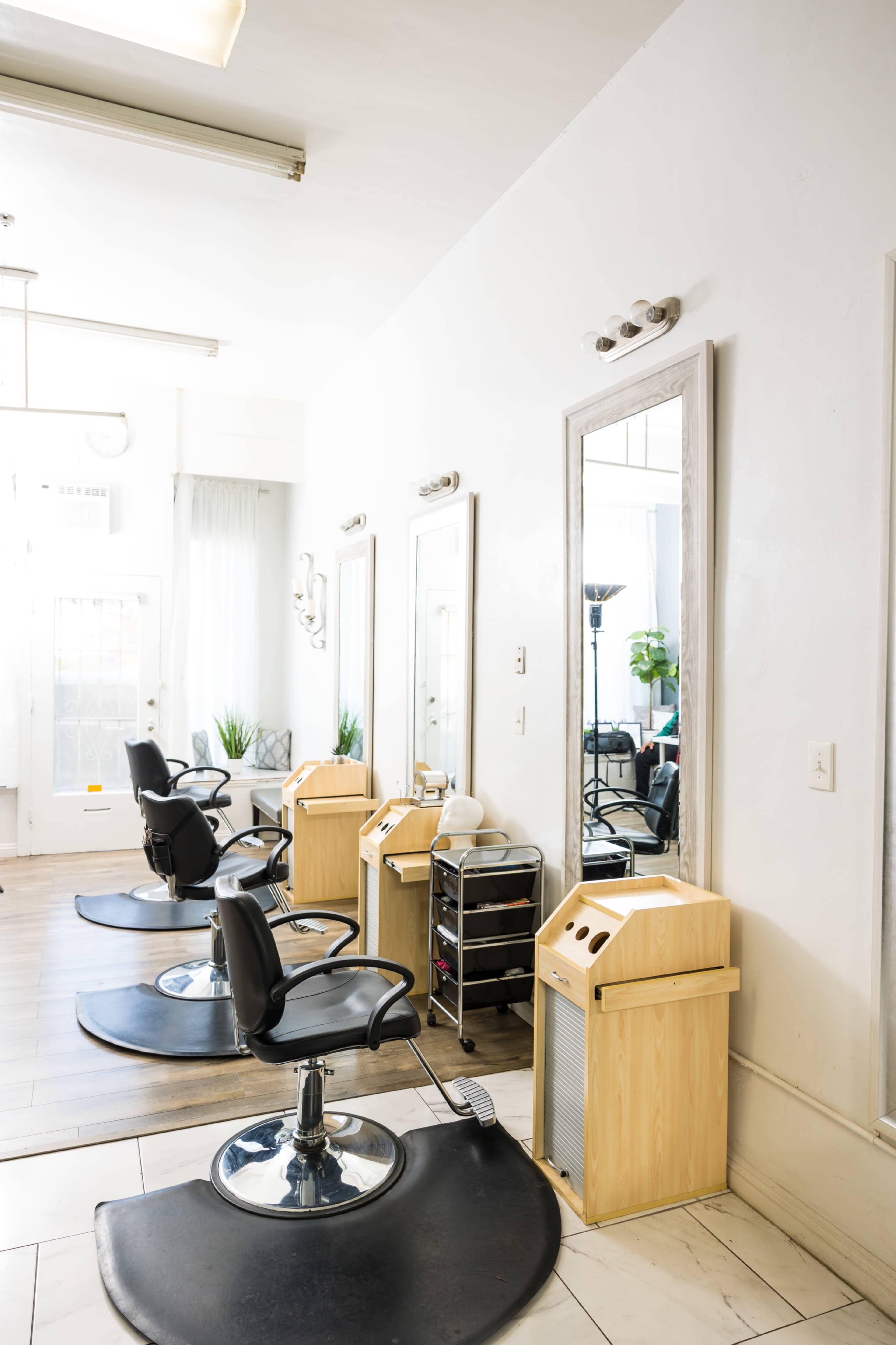 The image shows a bright hair salon featuring several black salon chairs, mirrors, and wooden styling stations.