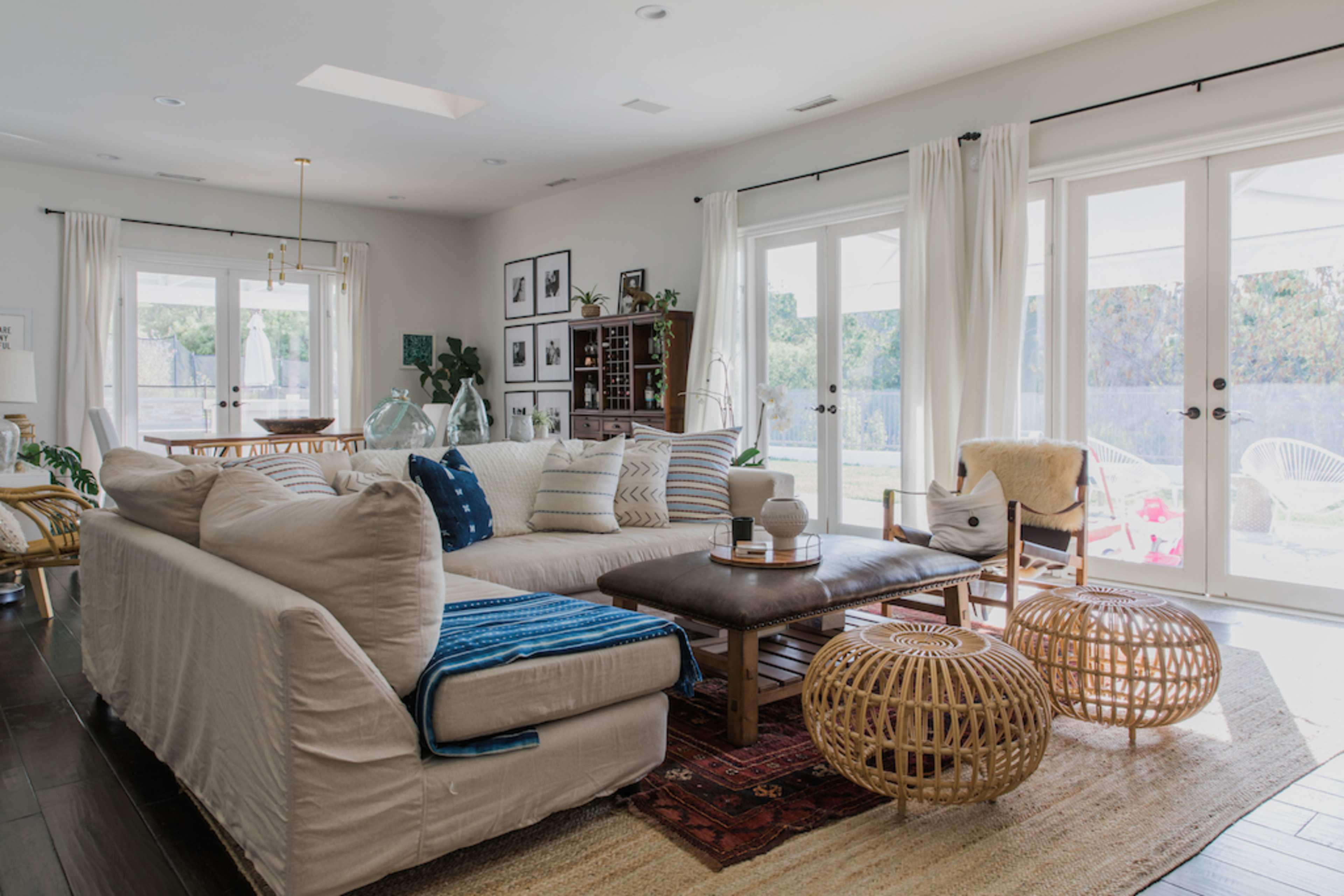 A spacious living room features a large beige sectional sofa, wooden coffee table, and decorative woven poufs, with natural light streaming in through sliding glass doors.