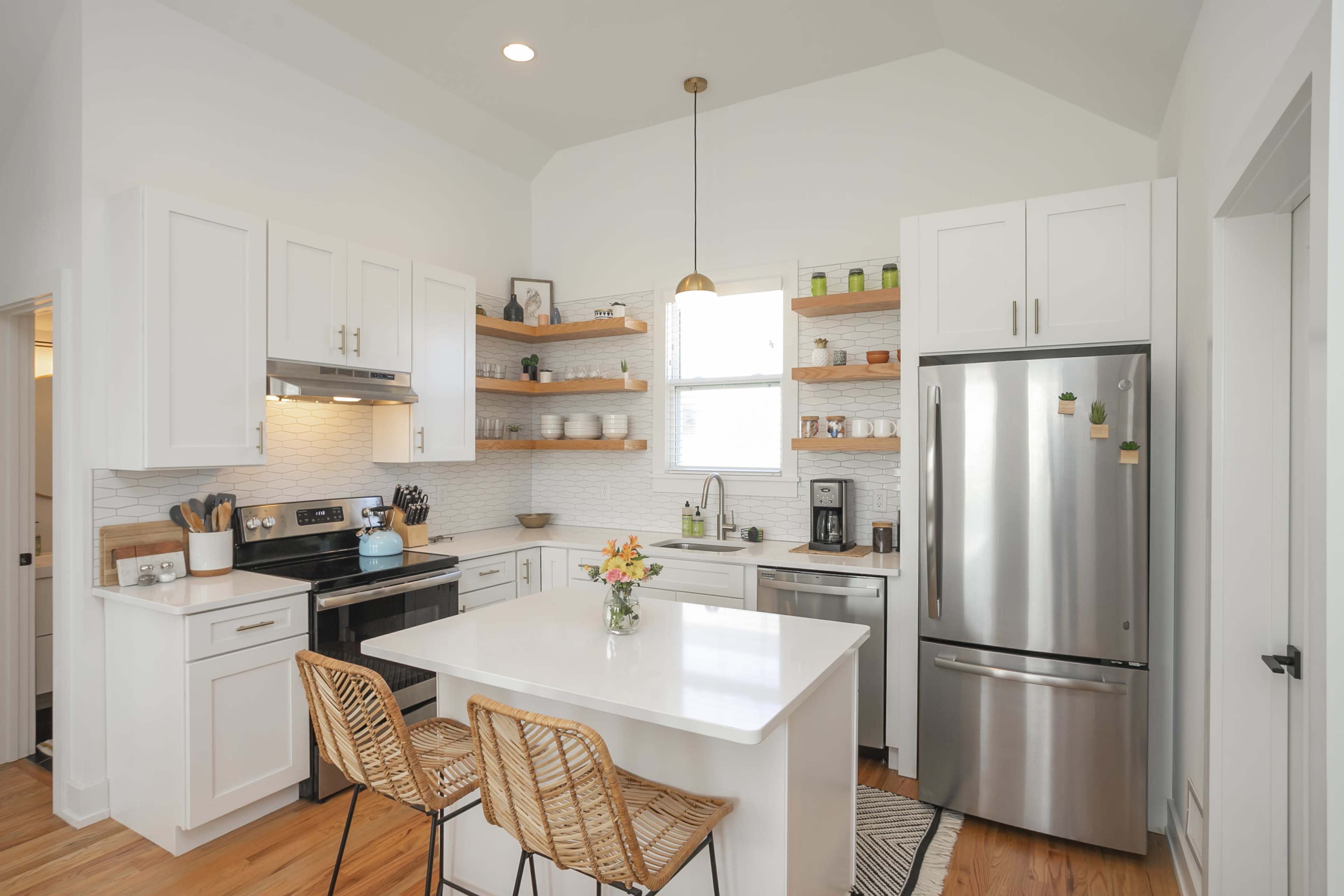 A modern kitchen features white cabinetry, stainless steel appliances, open shelving with various kitchen items, and a central island with two wicker chairs.