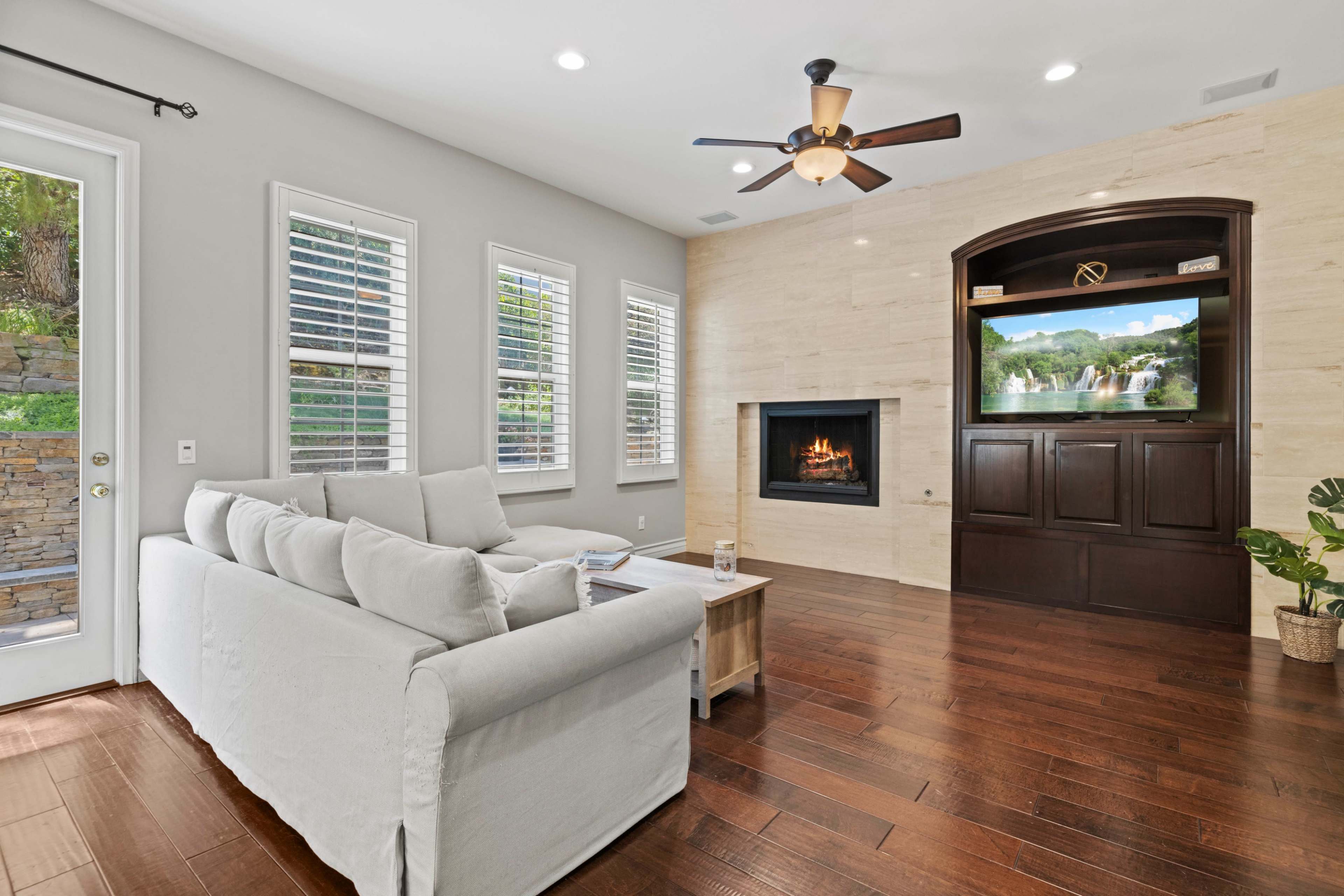 A modern living room features a light-colored sofa, hardwood flooring, a wall-mounted television, and a fireplace built into a stone panel backdrop.