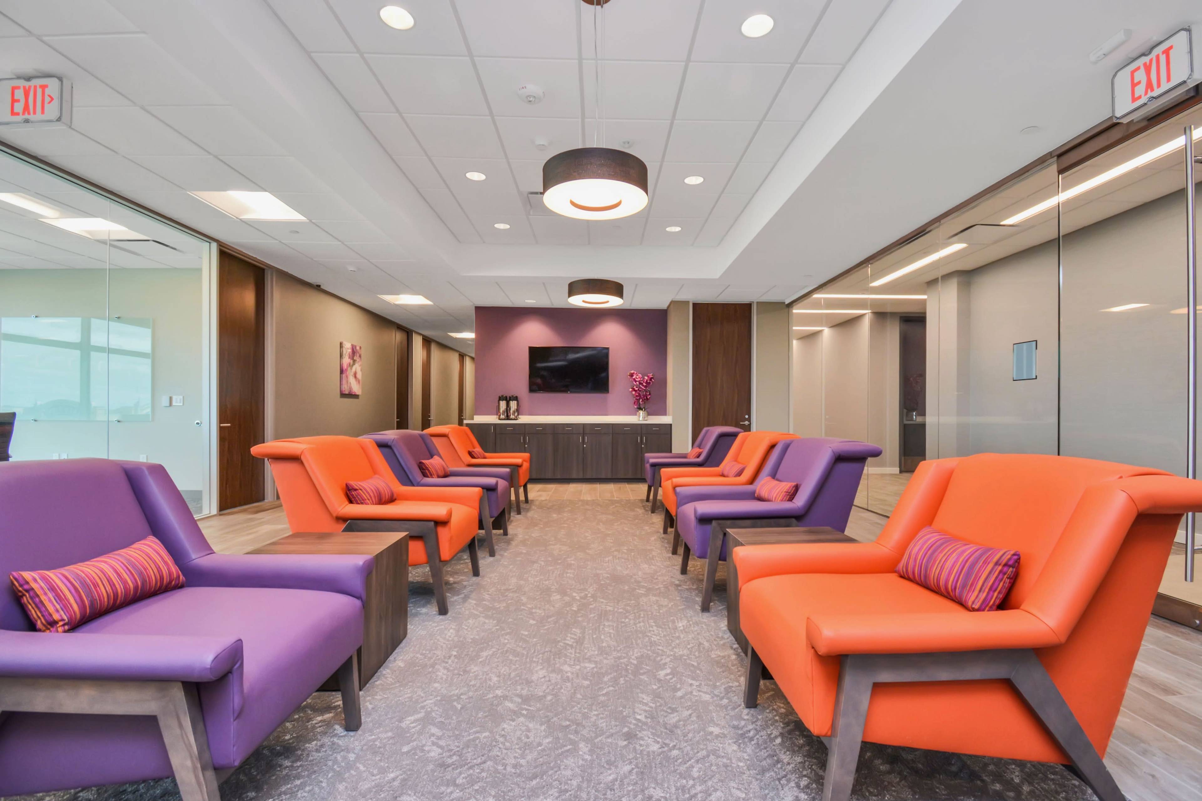 The image shows a modern waiting area furnished with purple and orange chairs arranged around a central space, featuring a flat-screen TV and decorative elements on the walls.