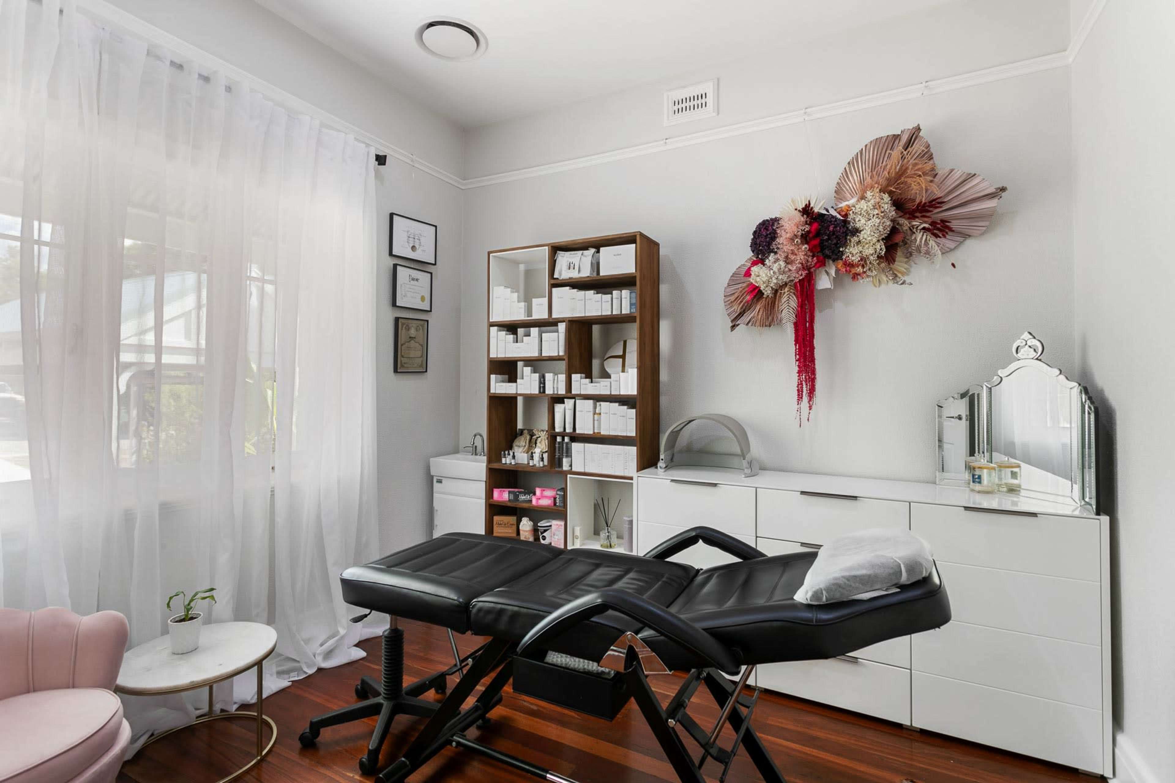 A beauty treatment room features a black treatment bed, a wooden shelving unit with neatly organized skincare products, and decorative floral arrangements on the wall.