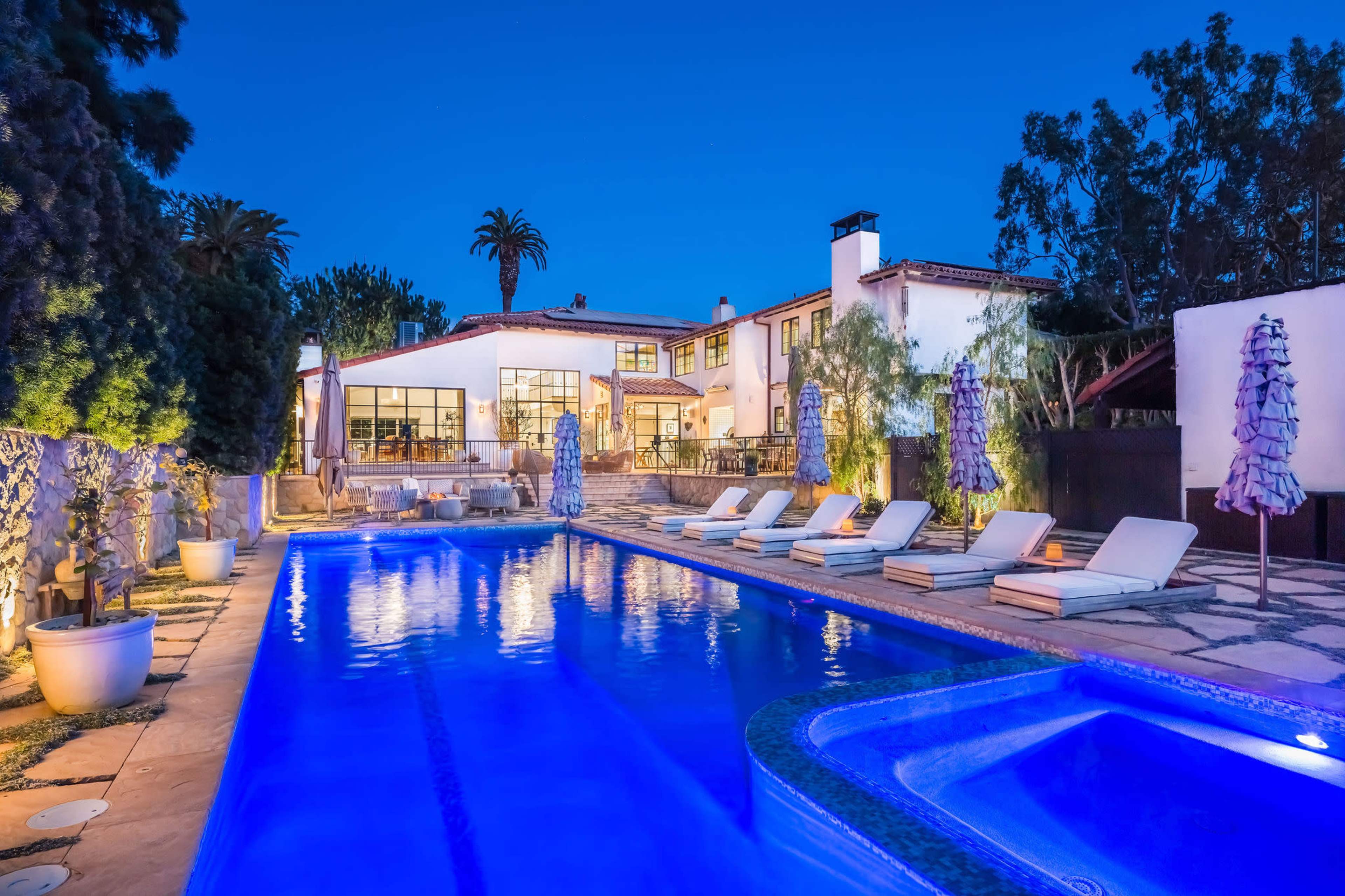 A large swimming pool with lounge chairs is set in a well-lit residential backyard, surrounded by lush greenery and a spacious house in the background.