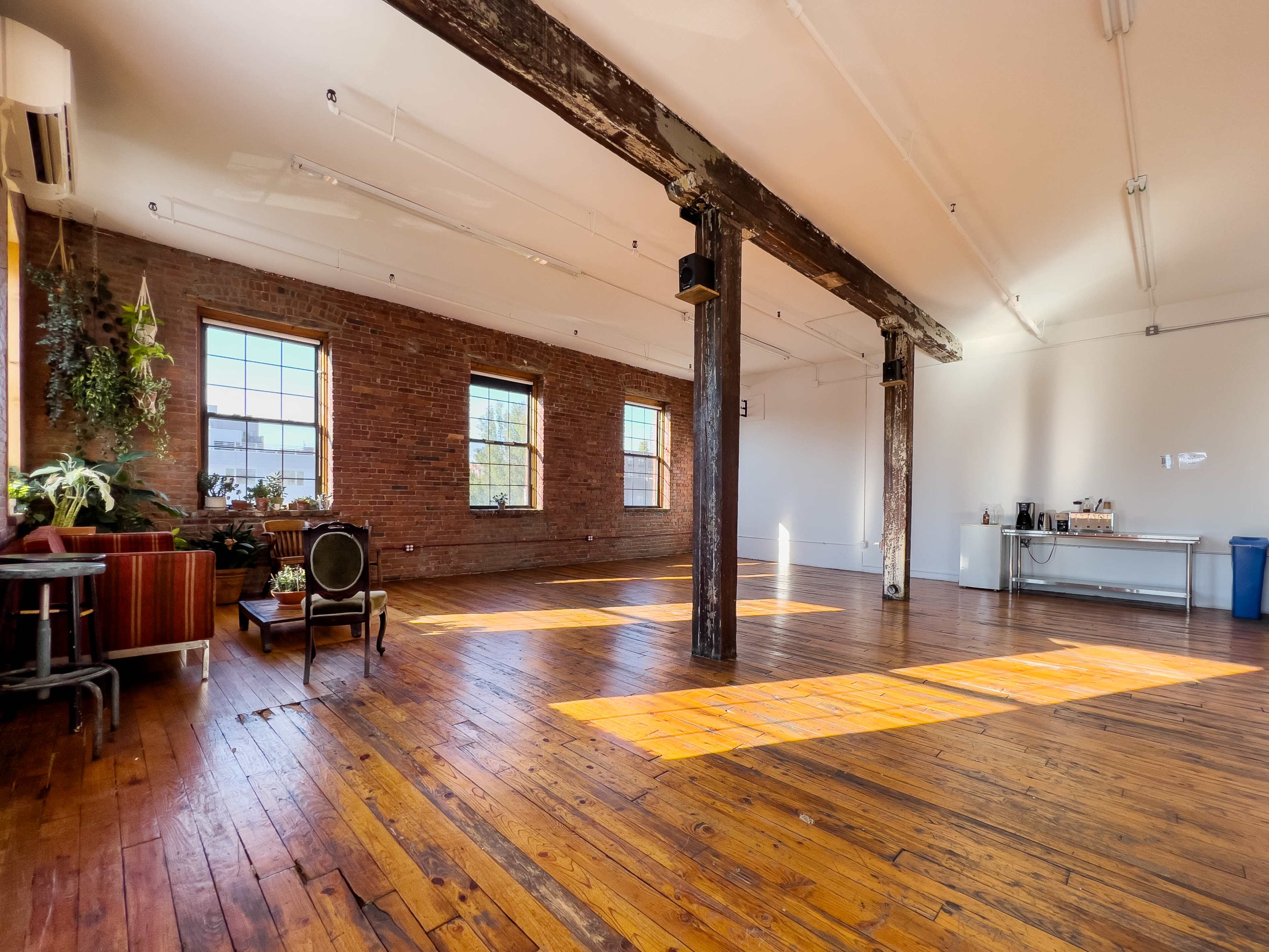 A spacious, well-lit room with hardwood floors, large windows, exposed brick walls, and wooden beams.