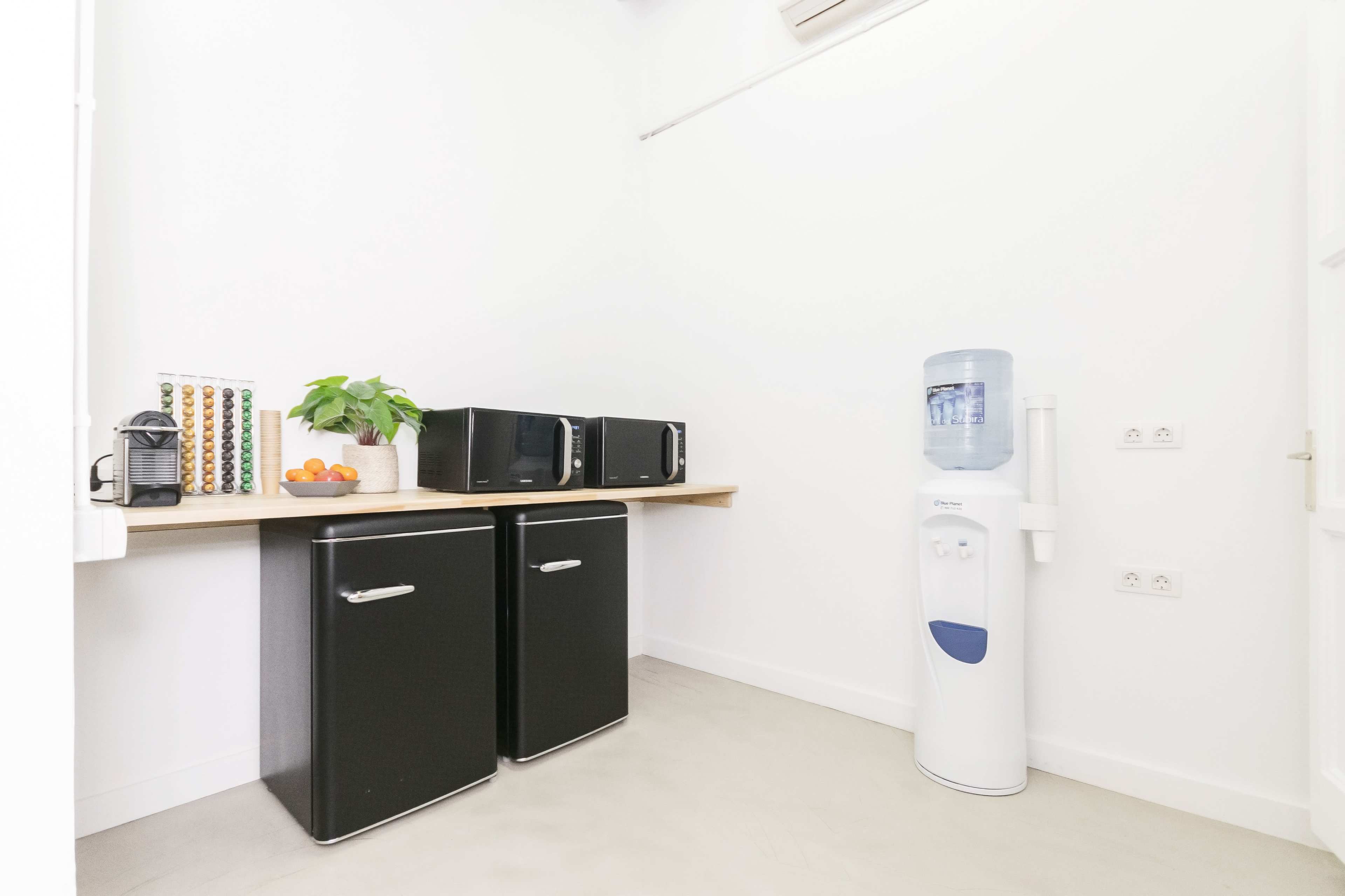 A small kitchen space featuring two black refrigerators, a countertop with a plant and fruit bowl, and a water cooler against the wall.