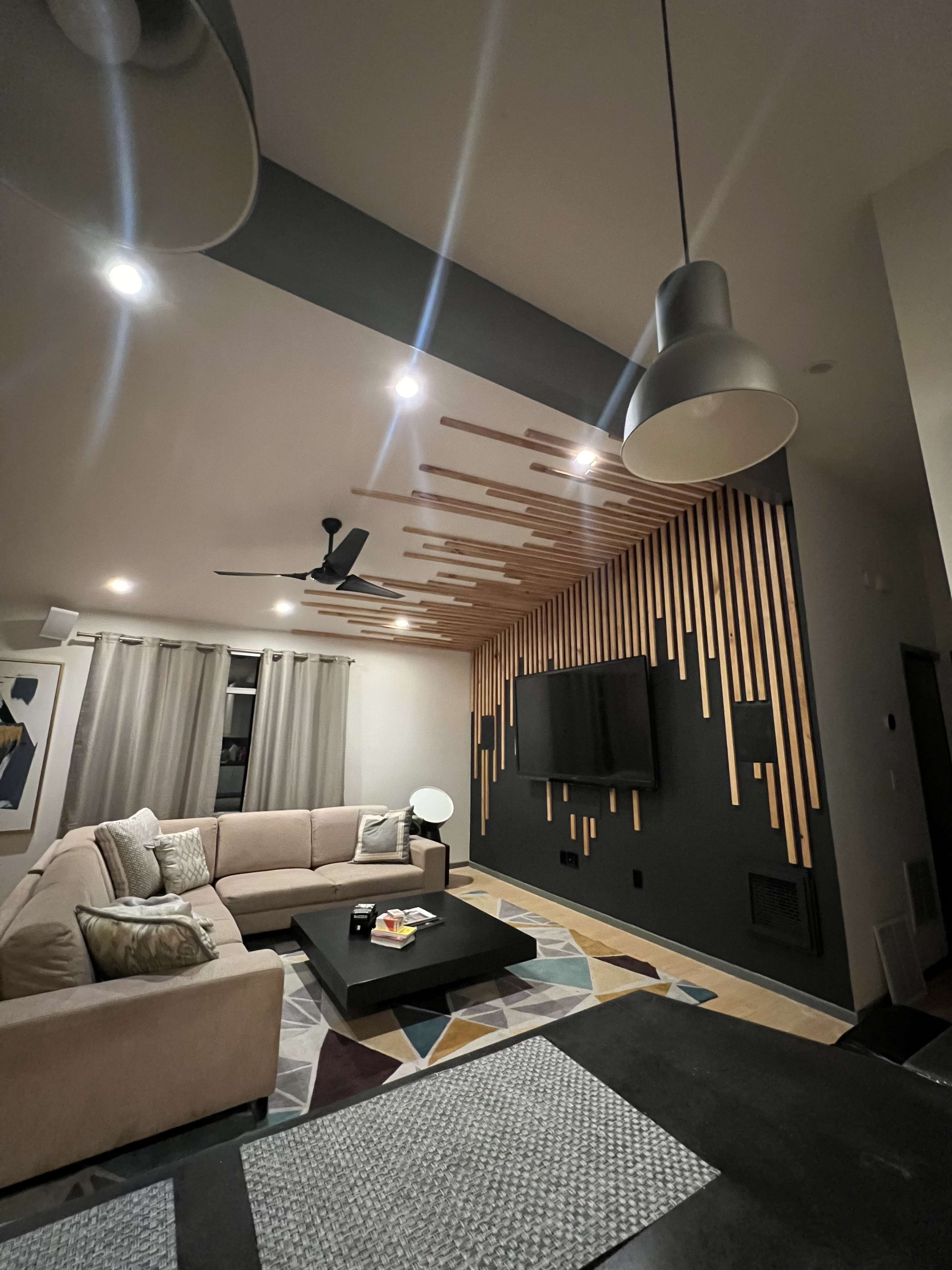 A modern living room features a large sectional sofa, a coffee table, a wall-mounted television, and decorative wood paneling along the ceiling.