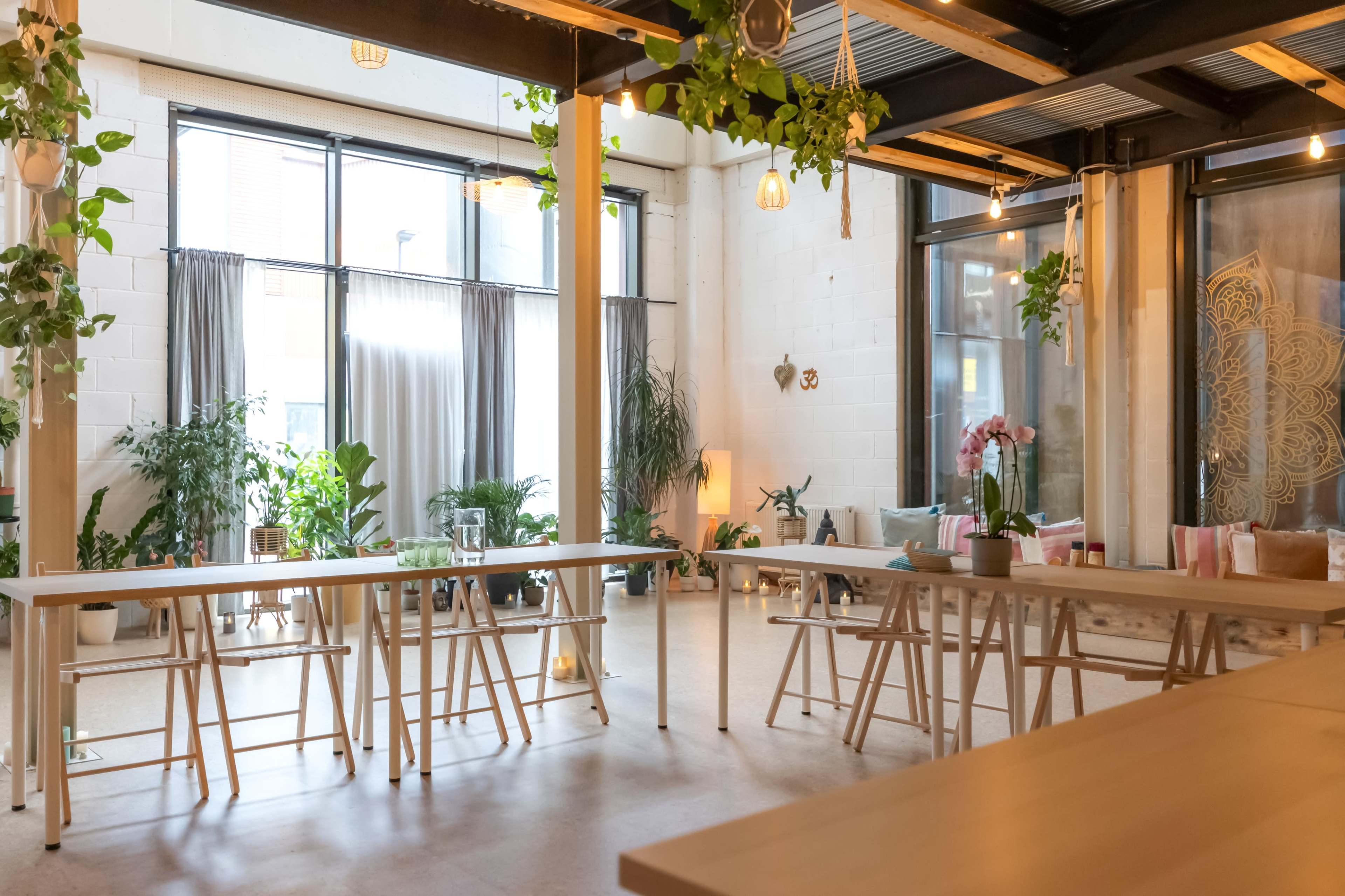 The image shows a bright, spacious room with large windows, wooden tables, and various potted plants scattered throughout.