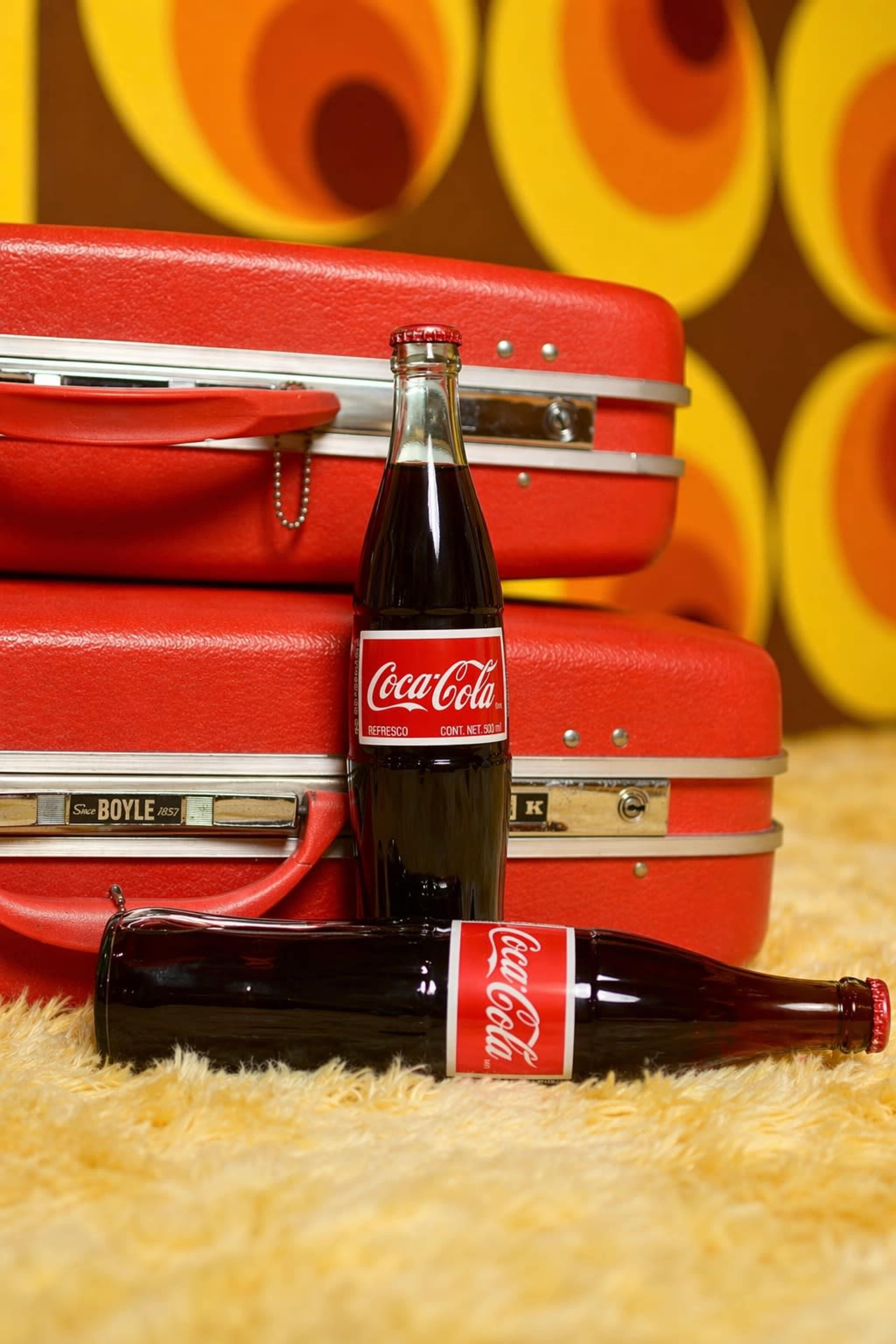 Two vintage Coca-Cola glass bottles are positioned near red suitcases against a backdrop of patterned yellow and orange wallpaper.