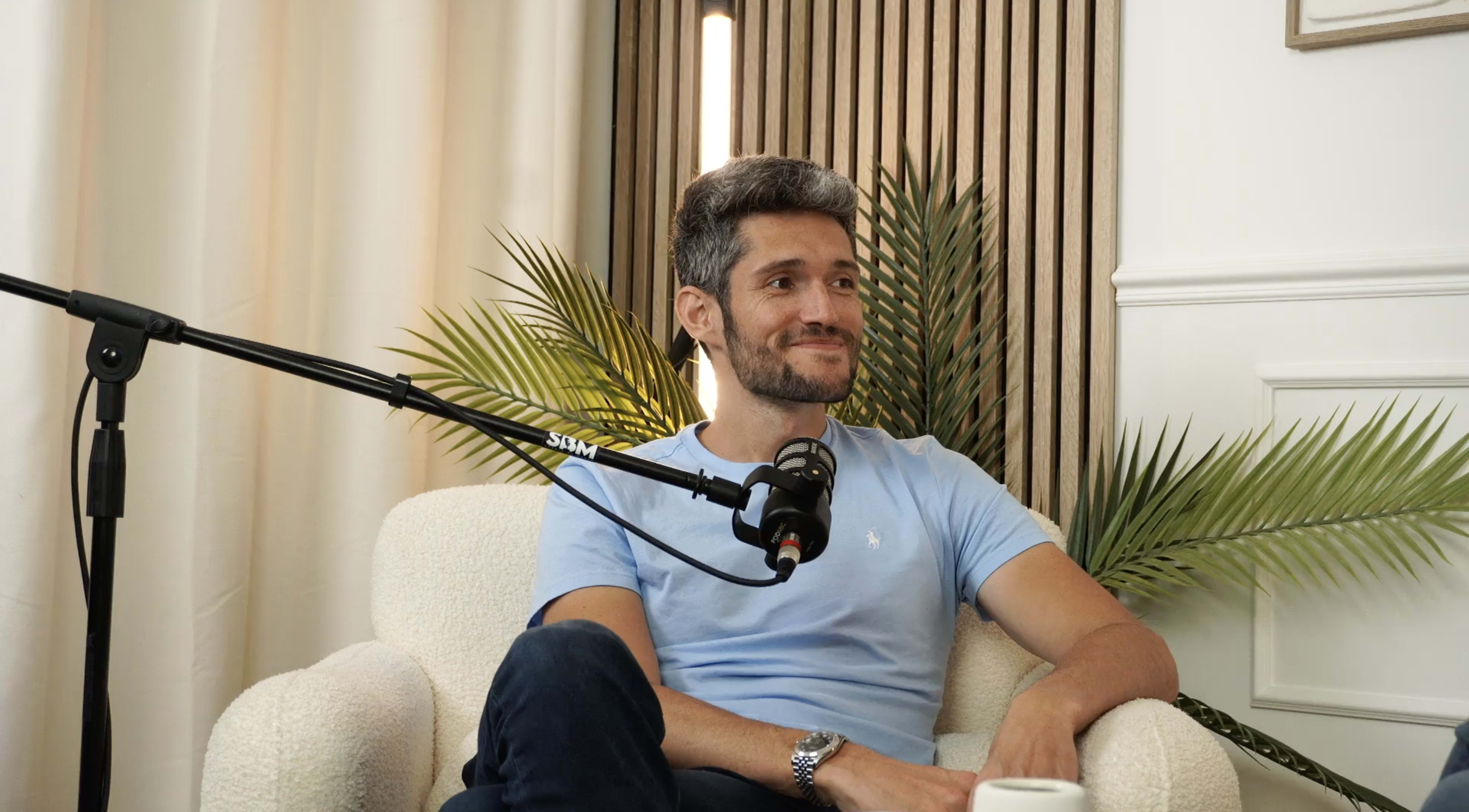 A man with short hair sits on a light-colored armchair, smiling while a microphone is positioned near him against a backdrop of indoor plants.