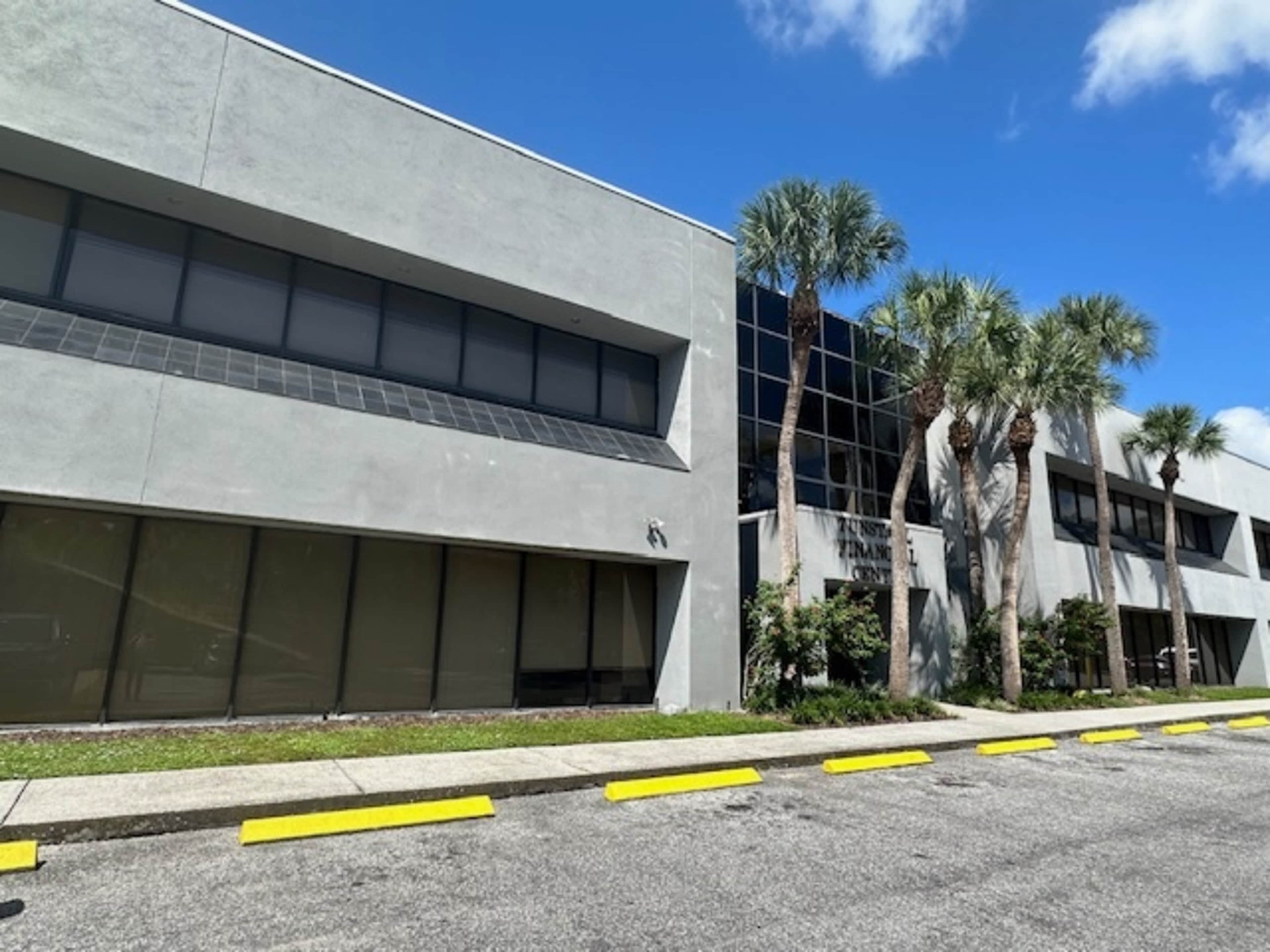 The image shows a modern gray building with multiple windows, palm trees along the front, and a clear blue sky.