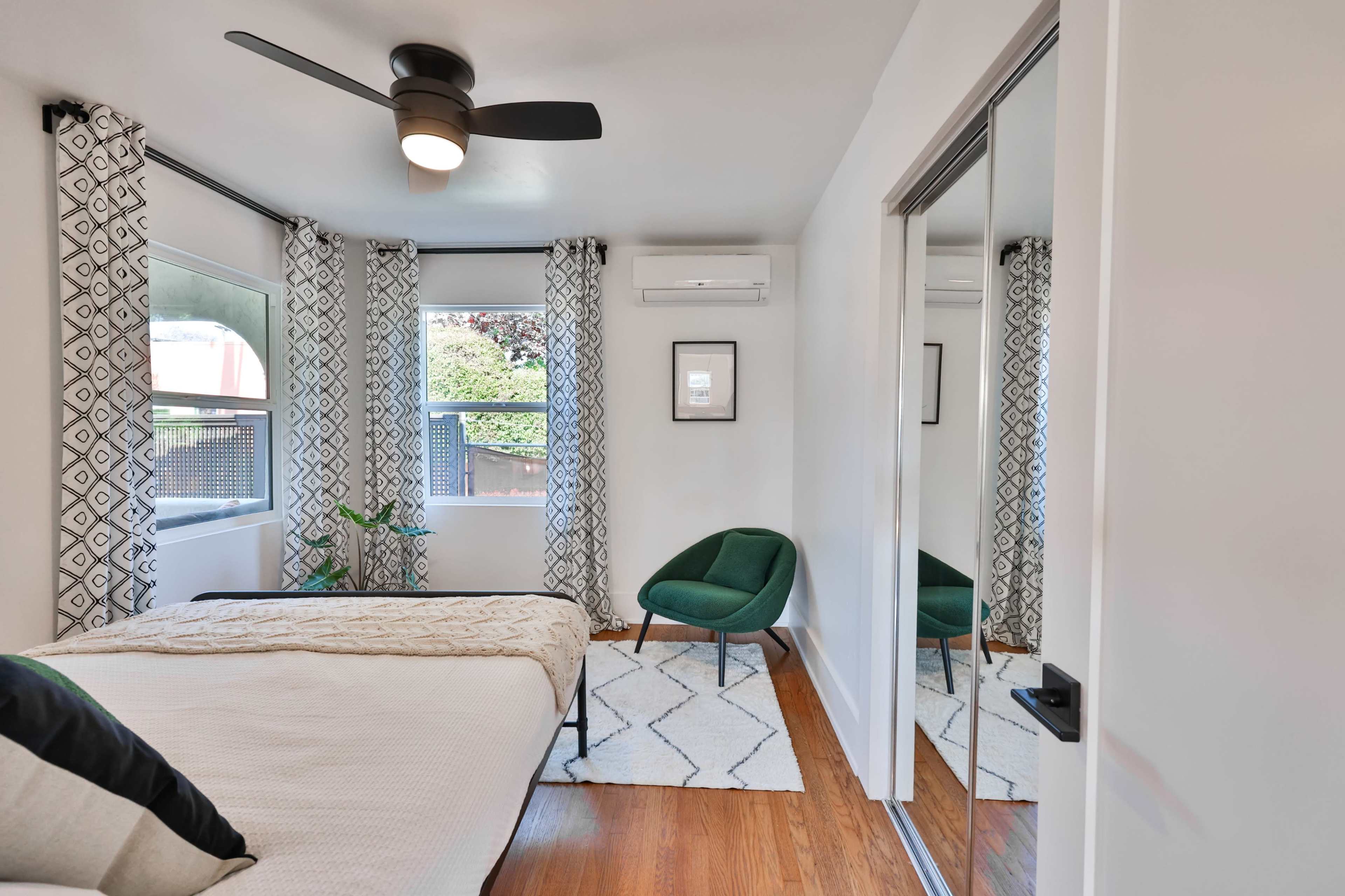 A bedroom with a bed, a green chair, and large windows covered by patterned curtains, along with a mirrored closet.